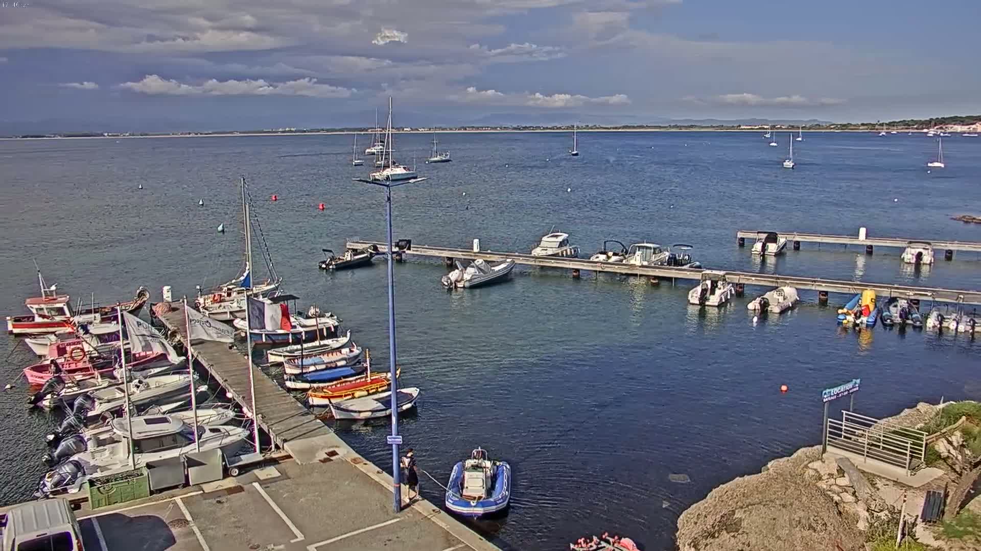 A partly cloudy day shows a marina filled with various boats and small vessels moored at docks extending into calm blue water.