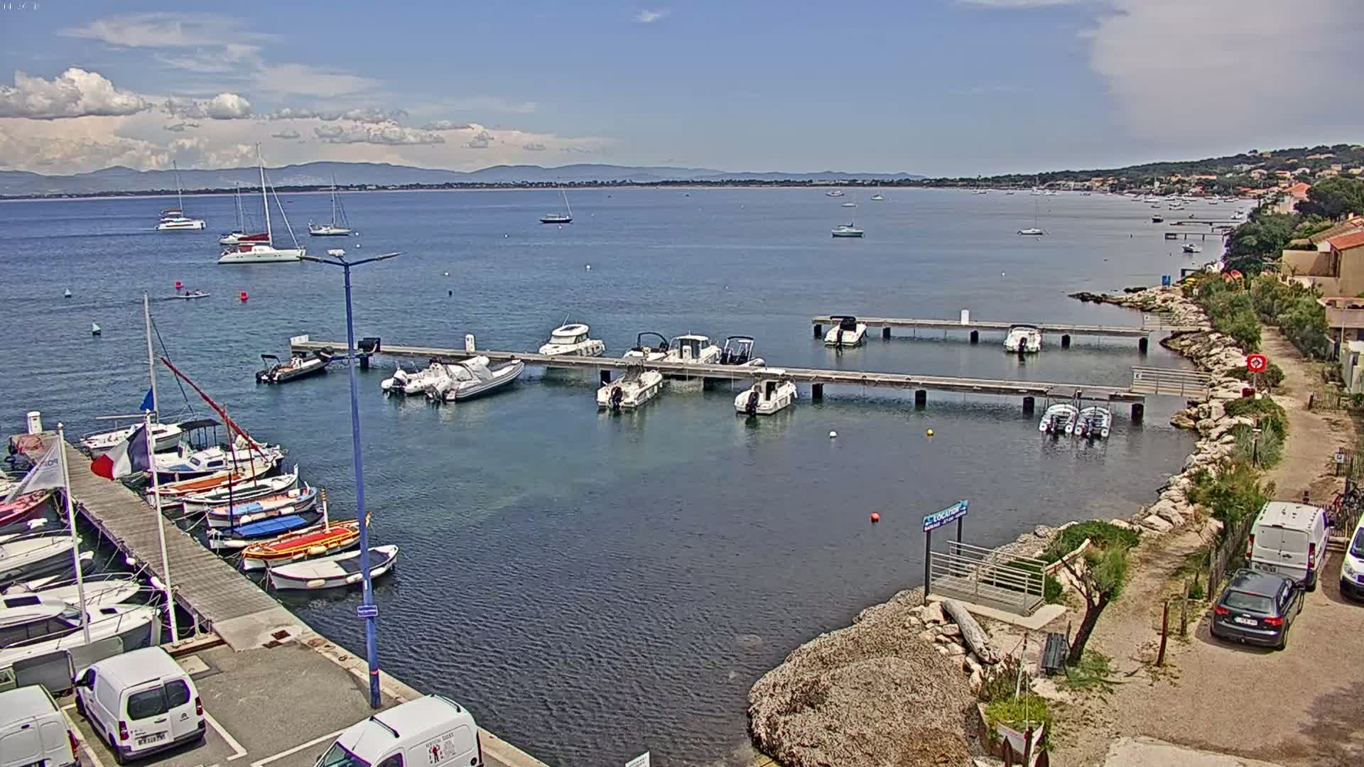 A sunny day at a harbor shows numerous boats docked at piers, with additional boats anchored in the calm water, and a distant shoreline visible under a partly cloudy sky.