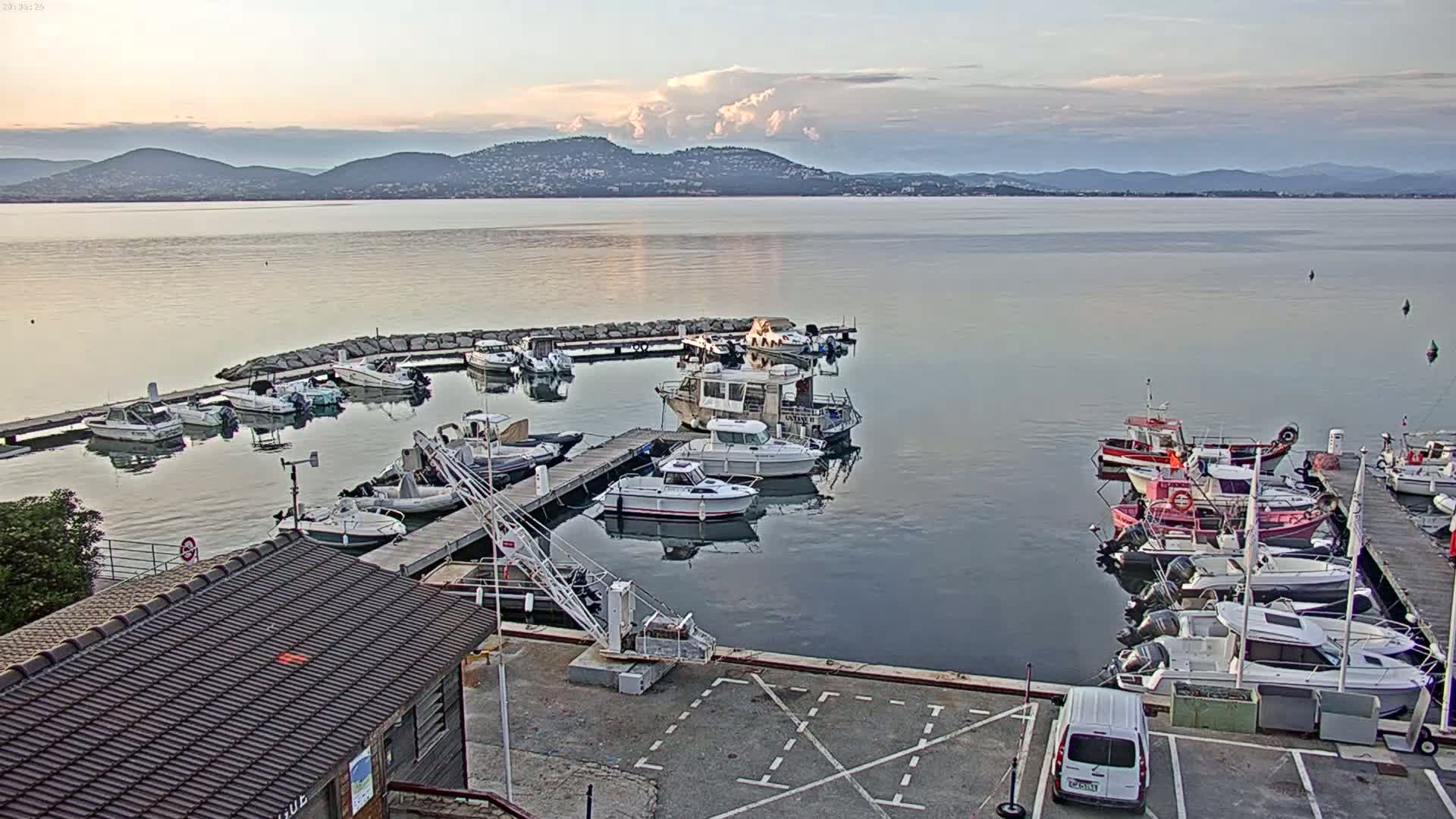 A calm body of water with many boats docked at a marina, under a partly cloudy sky with mountains in the distance.