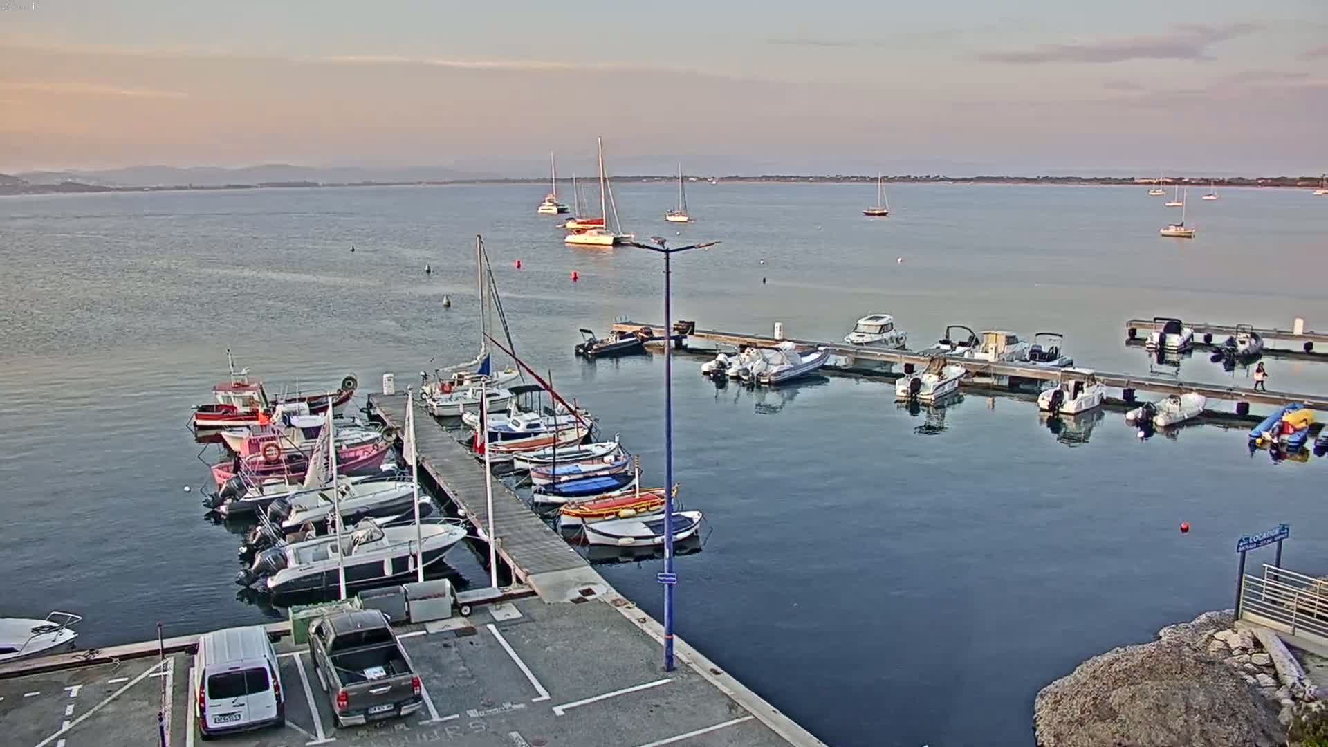 A calm, sun-dappled harbor filled with numerous boats docked at piers, with additional boats anchored in the calm water beyond.