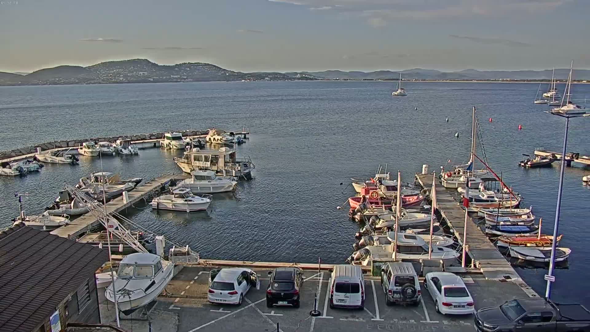 A marina filled with numerous boats of various sizes is situated near a parking lot, all under a mostly clear sky with distant mountains visible across calm water.