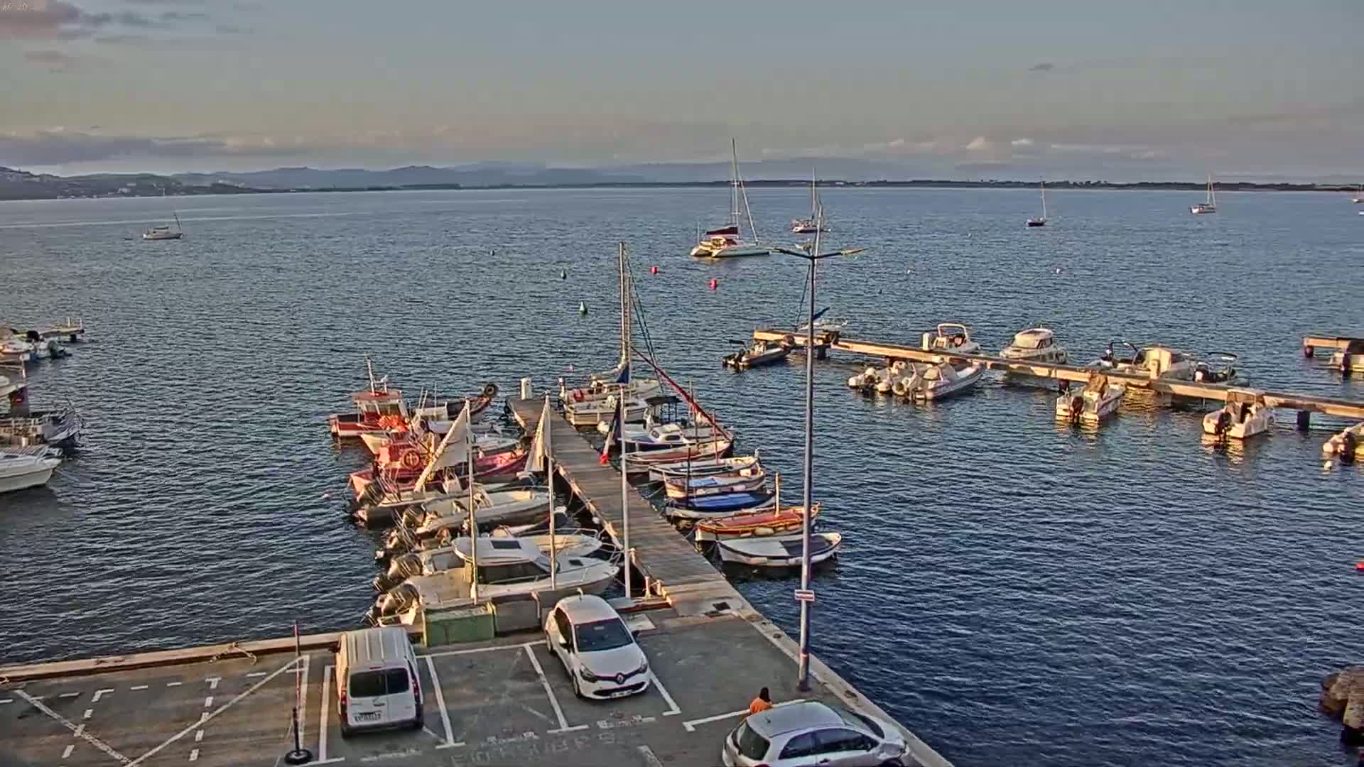 A calm body of water with numerous boats docked at a pier, several more boats anchored further out, and a few cars parked in a lot adjacent to the pier under a partly cloudy sky.