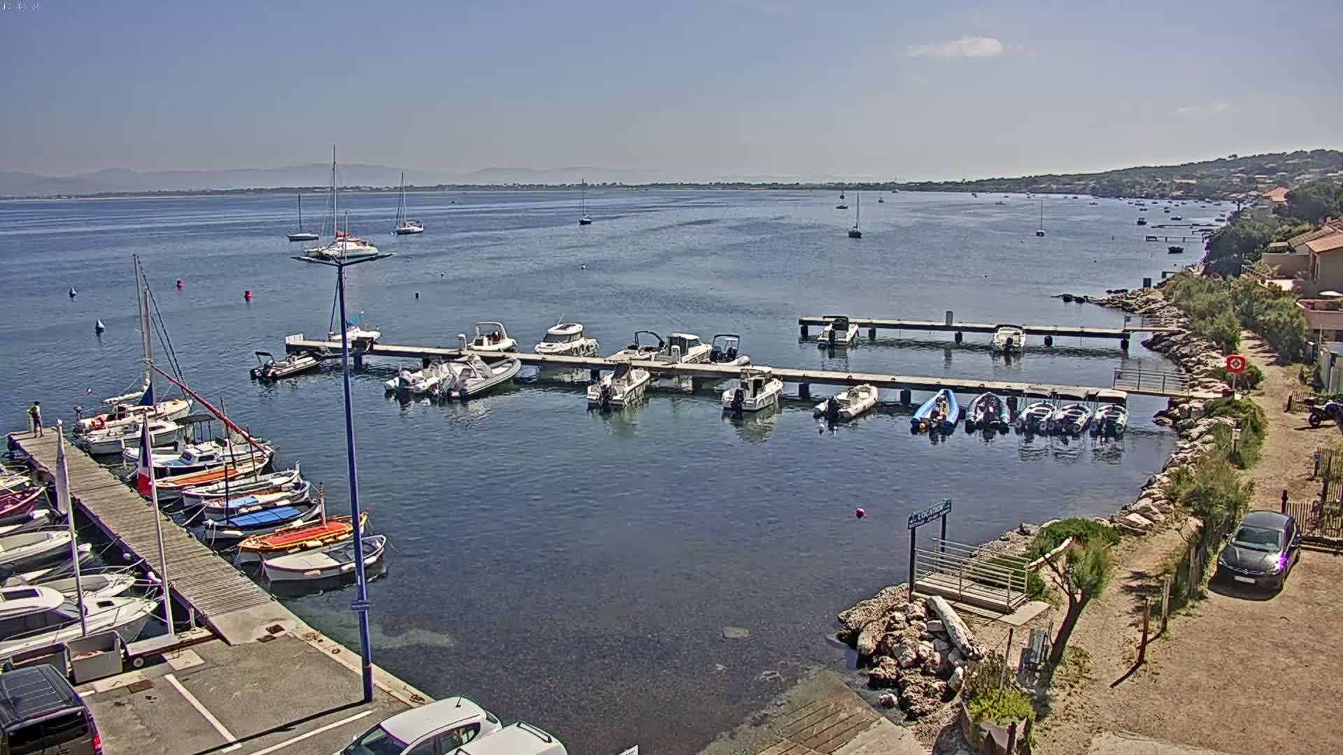A marina filled with numerous boats of various sizes is nestled on a calm, clear body of water under a sunny sky, with a distant shoreline visible in the background.