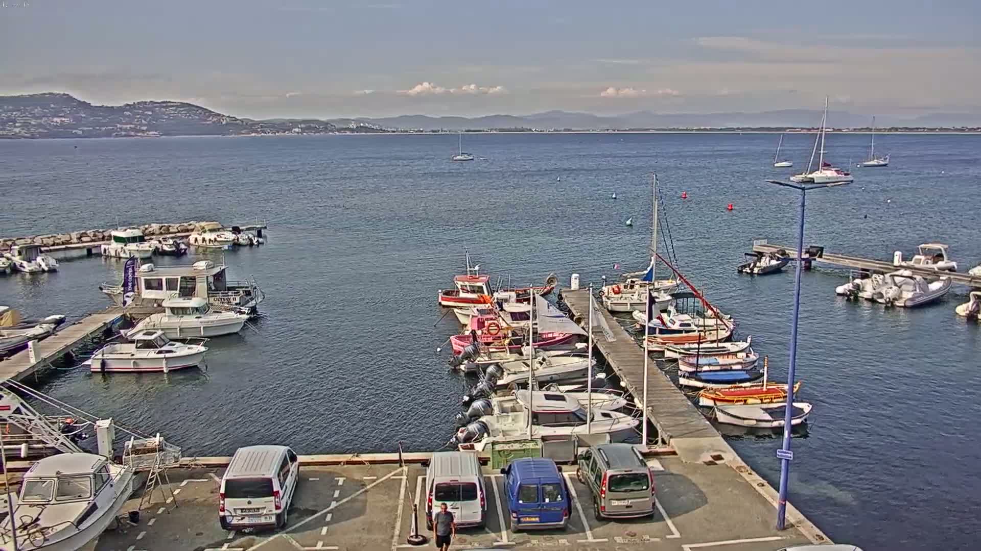 A marina filled with various boats and yachts is situated by a calm body of water under a partly cloudy sky, with a distant mountainous shoreline visible.