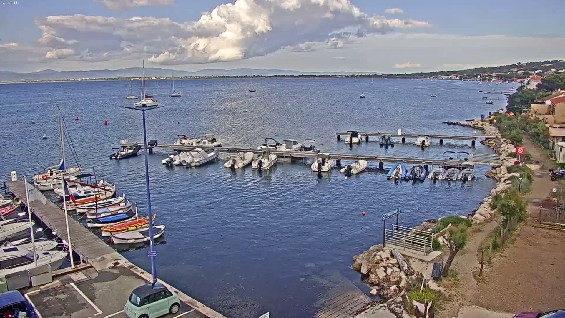 A partly cloudy day reveals a marina filled with boats docked along a pier, with additional boats anchored in the calm waters of a bay, beyond which lies a distant coastline under a partly cloudy sky.