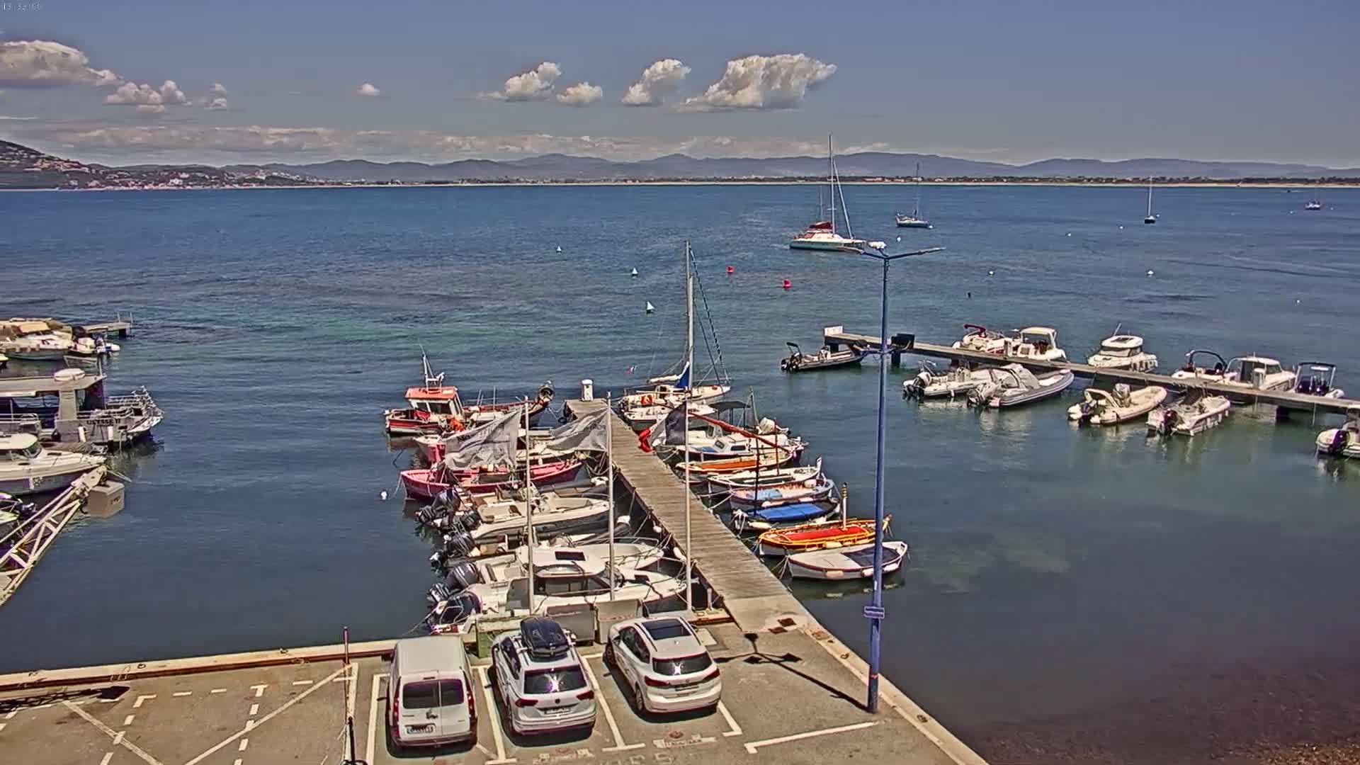 A sunny day shows a marina filled with sailboats and motorboats docked along a pier, with additional boats moored in the water beyond the pier, and a distant coastline visible across a calm bay.