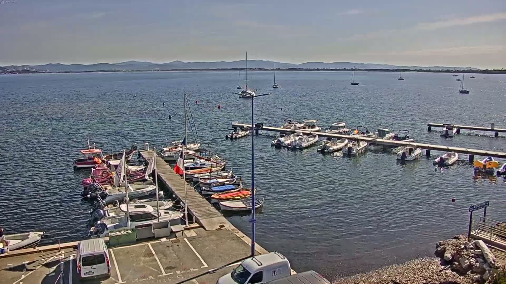 Numerous boats are docked at a marina on a calm, sunny day, with distant mountains visible across the water.