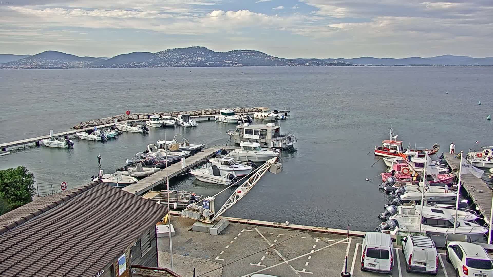 A marina filled with numerous boats of various sizes is situated near a calm body of water with mountains visible in the distance under a partly cloudy sky.