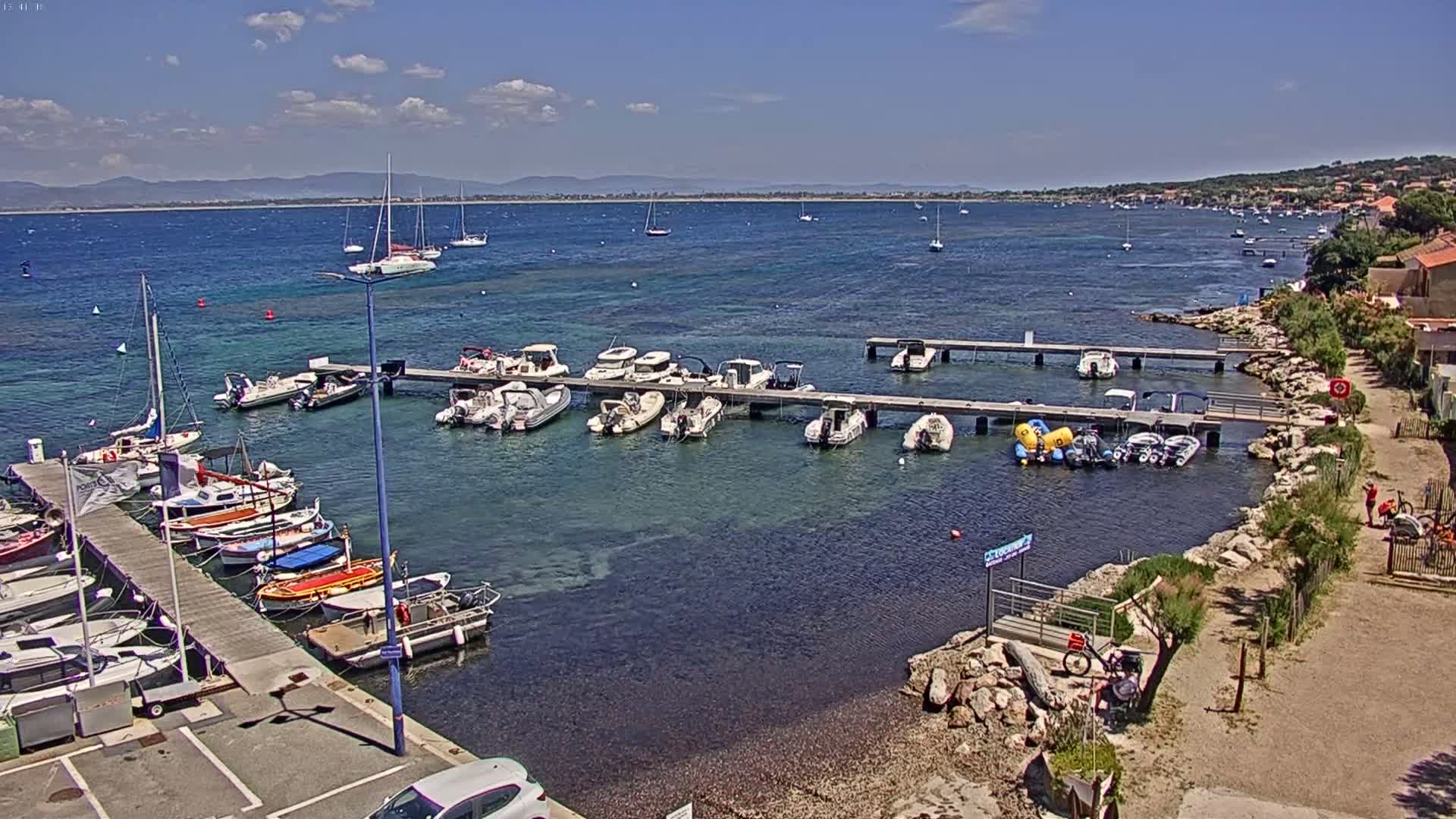 A sunny day shows numerous boats docked at a marina, with a calm, clear body of water extending to a distant shoreline.