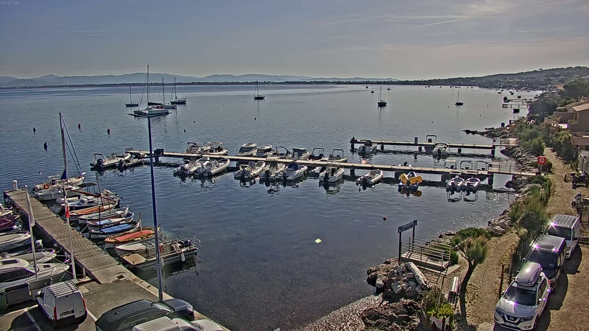 A calm bay with numerous boats docked at piers, under a clear, sunny sky, with distant mountains visible on the horizon.