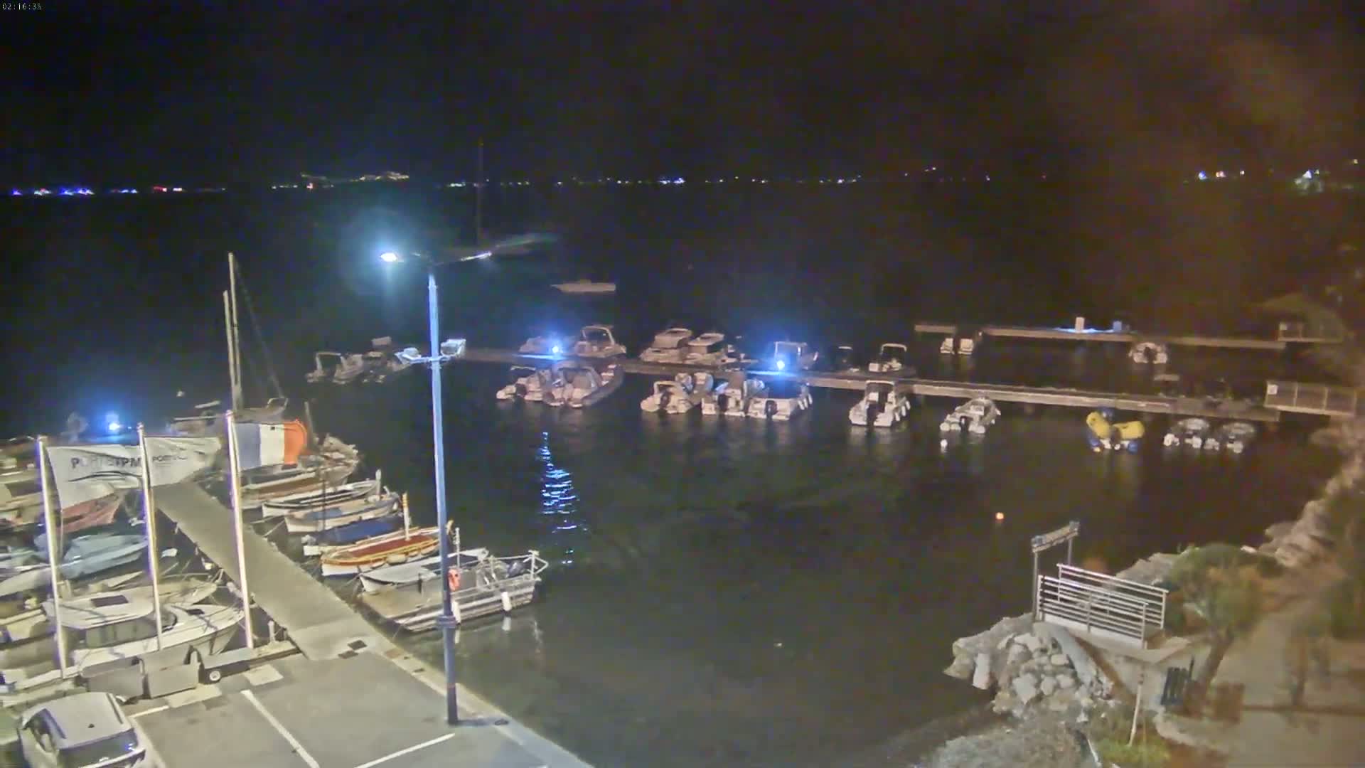 At night, numerous boats are moored at a dock along a calm body of water.