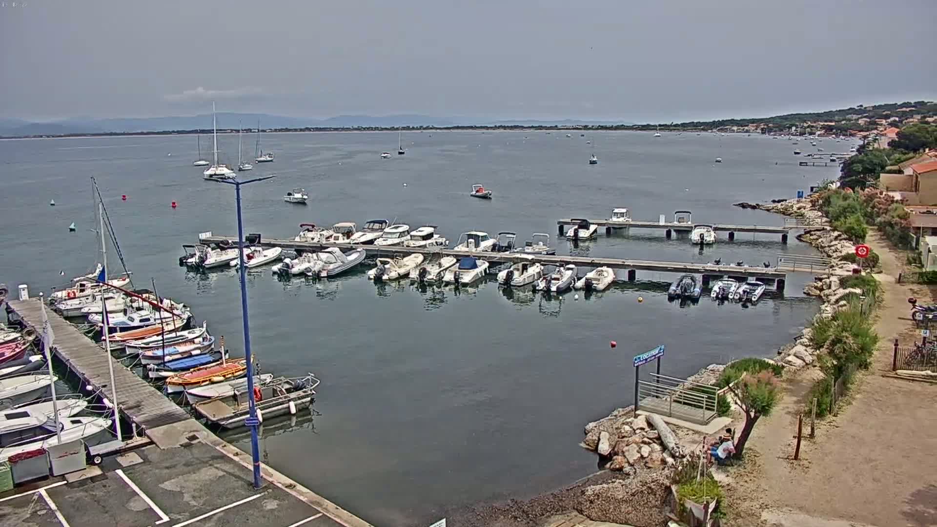 A marina filled with numerous boats of varying sizes is situated on calm, gray water under an overcast sky, with a distant shoreline visible in the background.