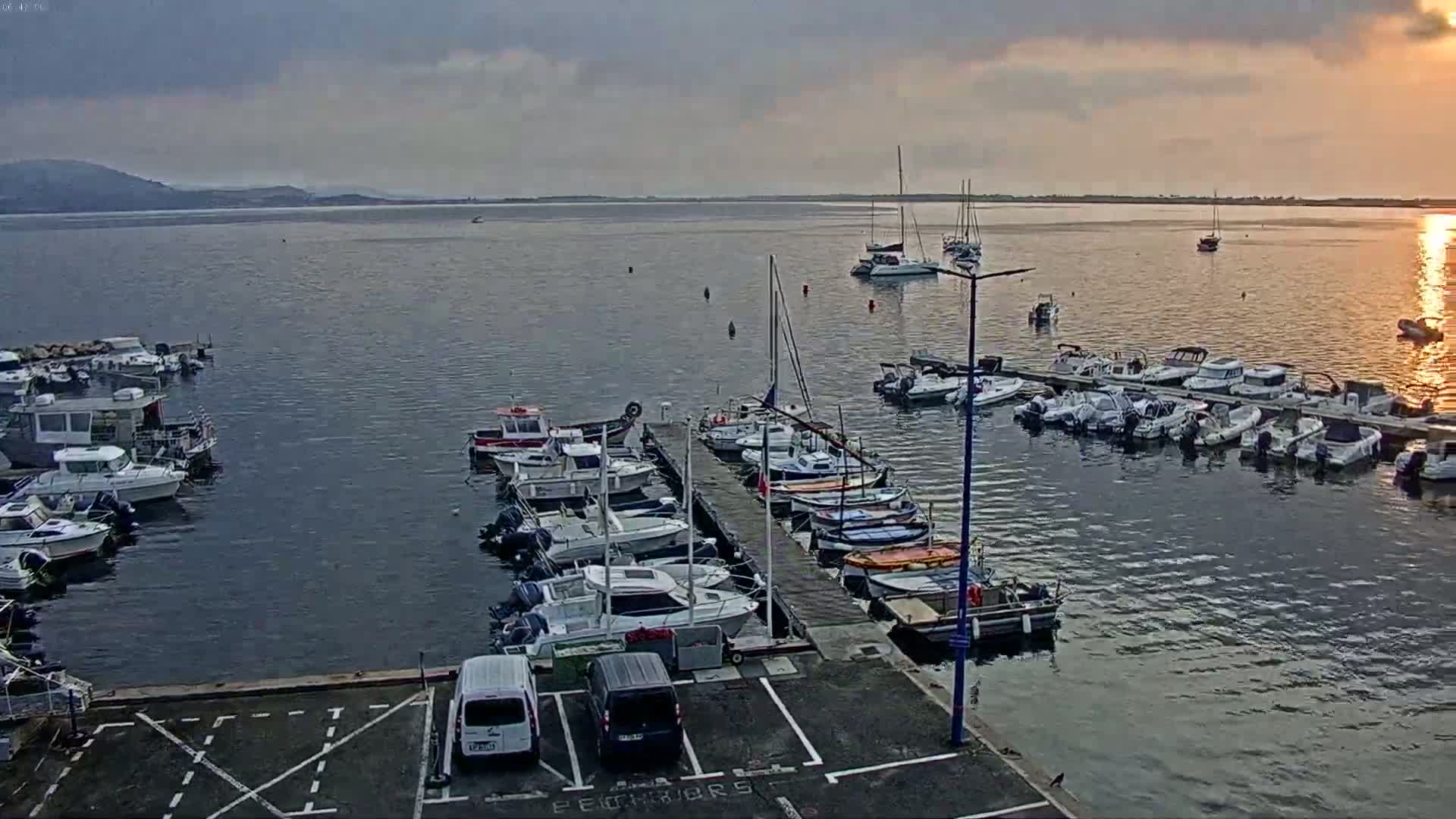Numerous boats are docked at a marina on calm water under a partly cloudy sky, with a few sailboats visible further out in the bay.