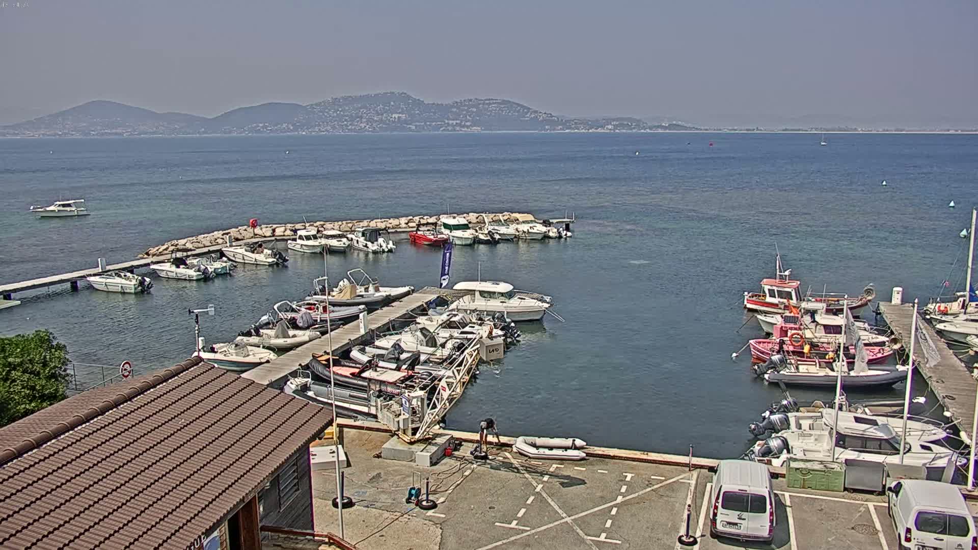 A marina filled with numerous boats of varying sizes is situated on calm, clear water, with a mountain range visible in the distance under a bright, sunny sky.