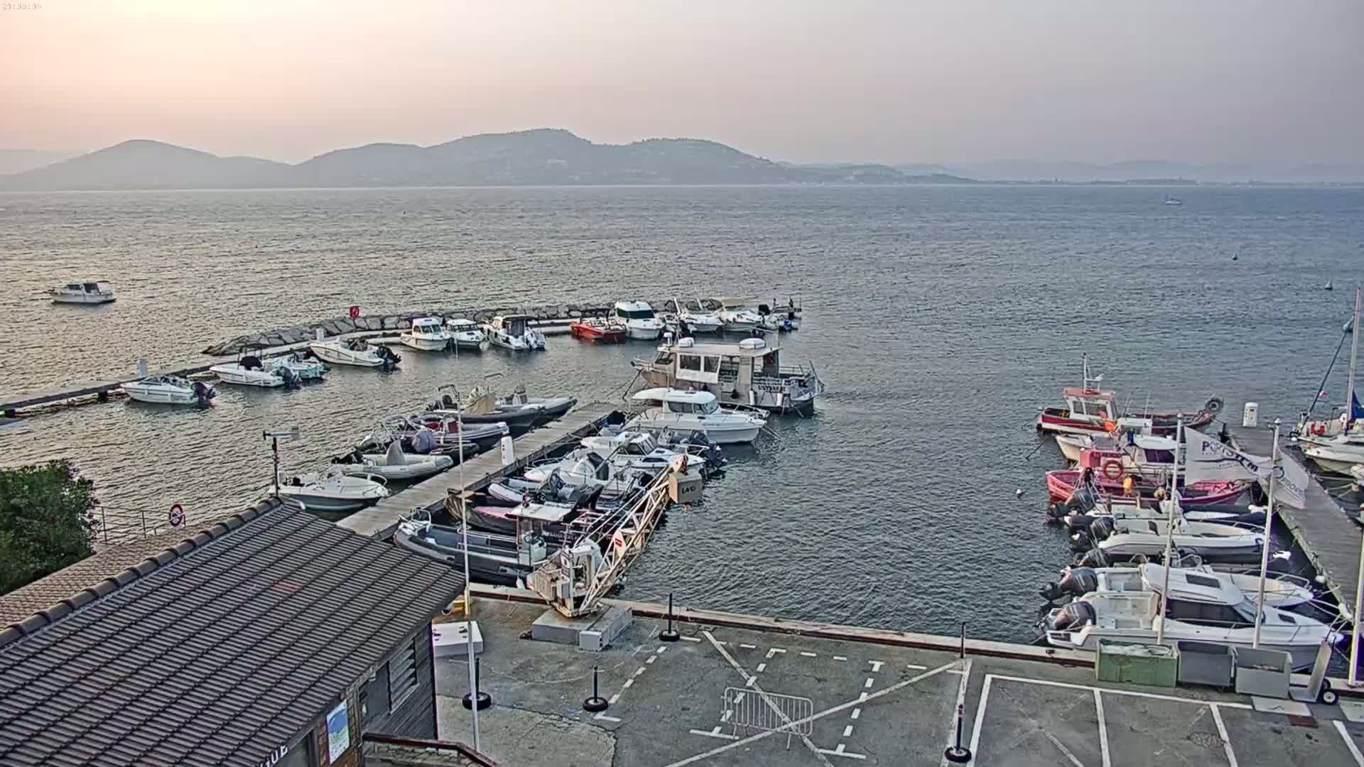 A marina filled with numerous boats of varying sizes is situated on calm water, with a hazy mountain range visible in the distance under a muted, overcast sky.