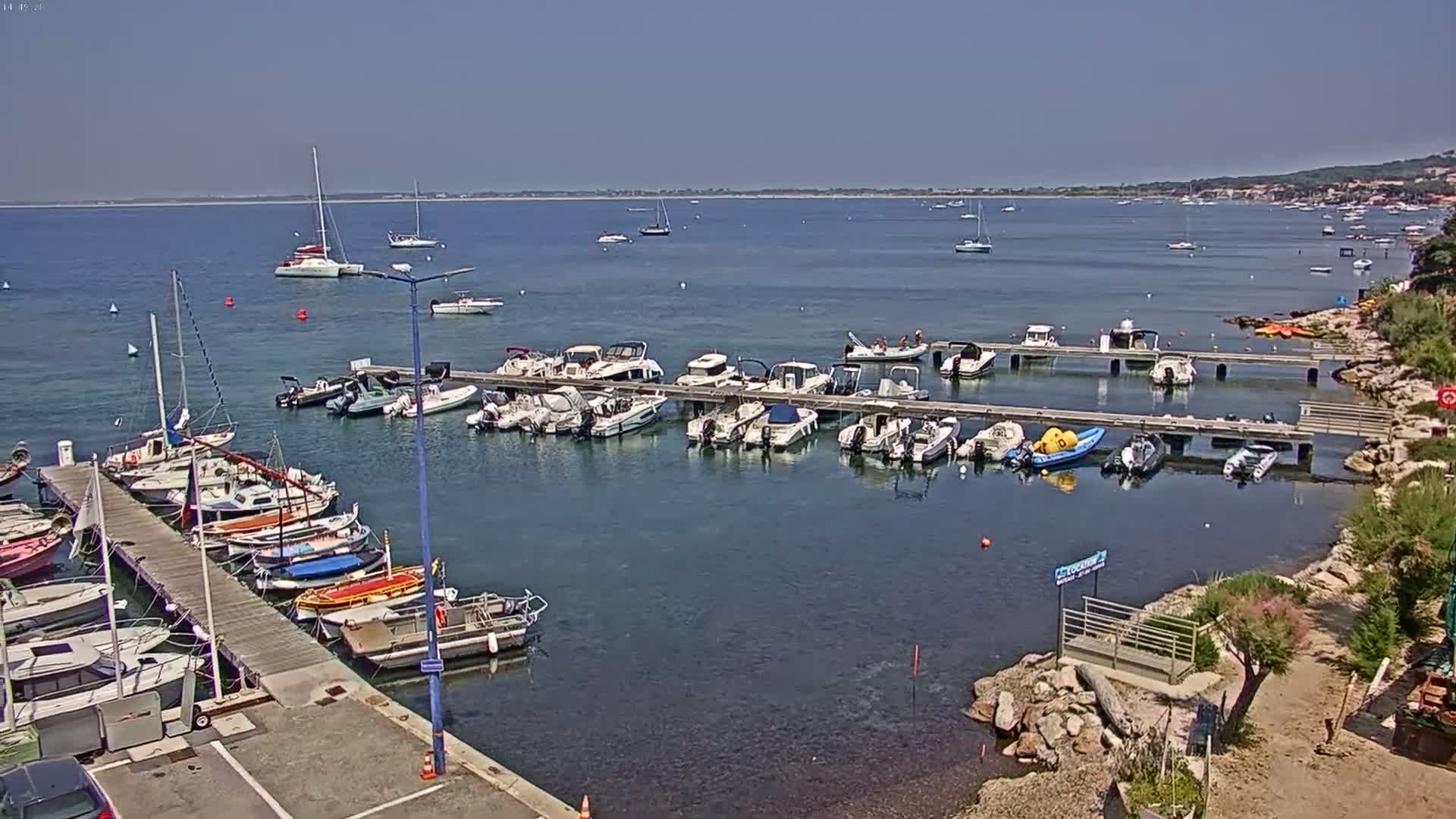 A marina filled with numerous sailboats and motorboats is situated on calm, clear water under a bright, sunny sky.