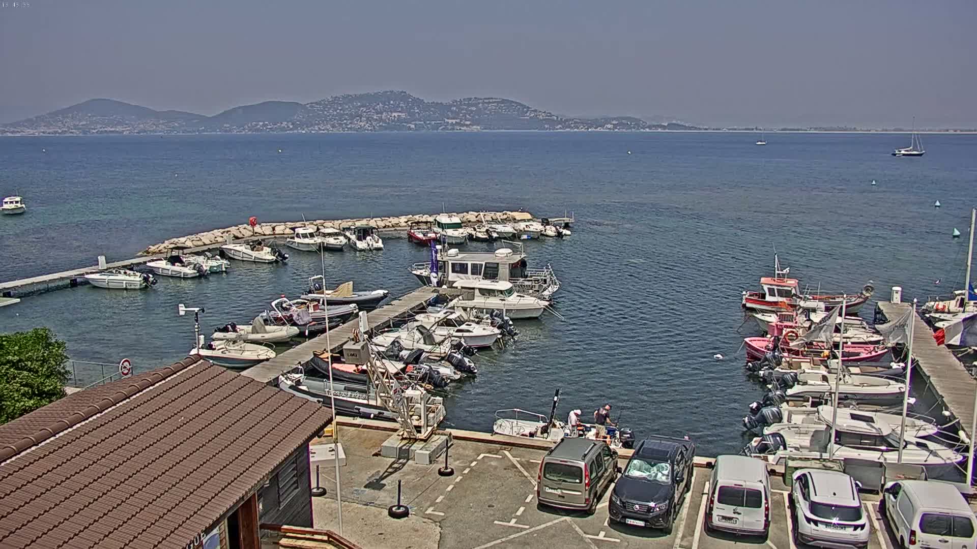 A marina filled with various sized boats and yachts is situated on calm, blue water next to a parking lot under a clear, sunny sky with distant mountains visible on the horizon.