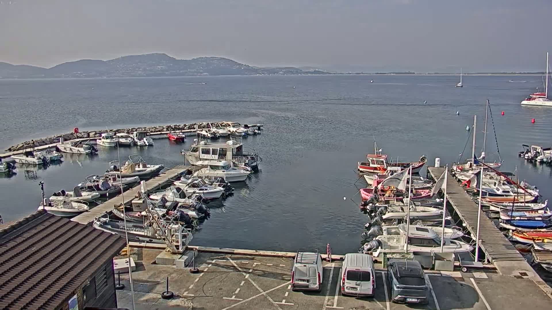 A marina filled with numerous boats of varying sizes is situated on calm water under a hazy sky, with distant mountains visible in the background.