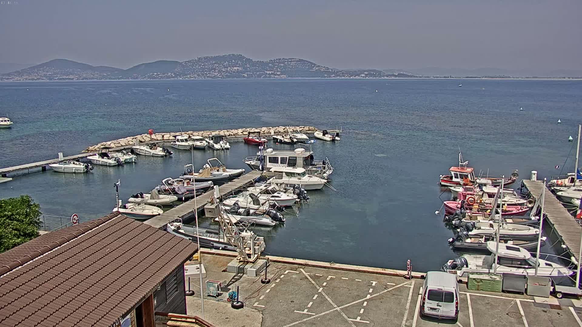 A marina filled with numerous boats of various sizes is situated near a body of water under a clear, sunny sky with mountains visible in the distance.