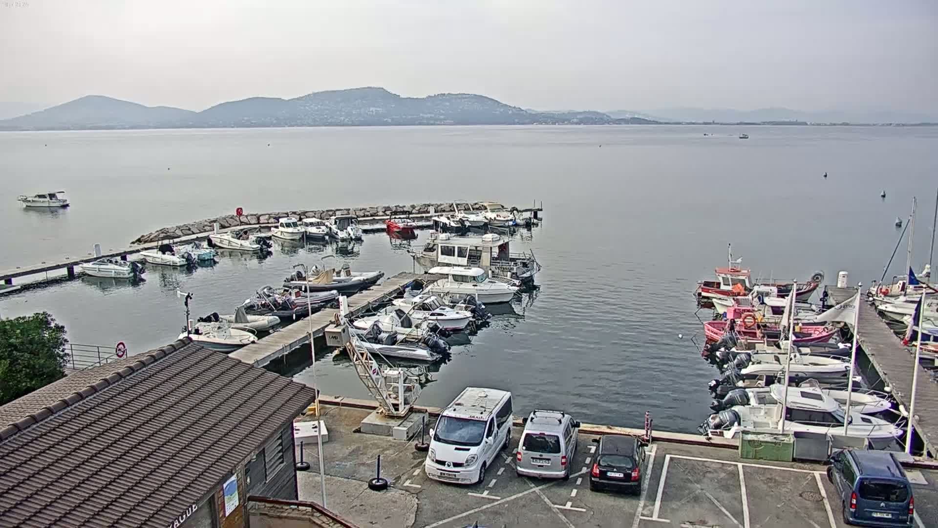 A marina filled with various sized boats and yachts is situated on calm water, with a hazy mountain range visible in the background under a cloudy sky.