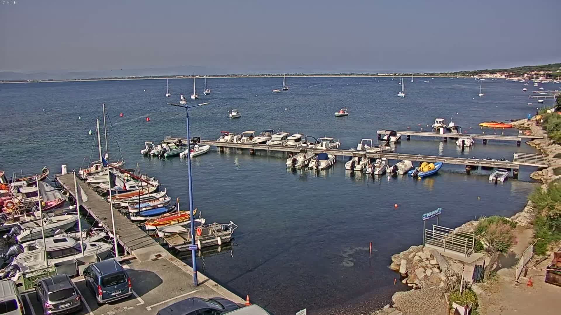 A sunny day at a marina shows numerous boats docked at piers, with additional boats anchored in the calm water beyond.