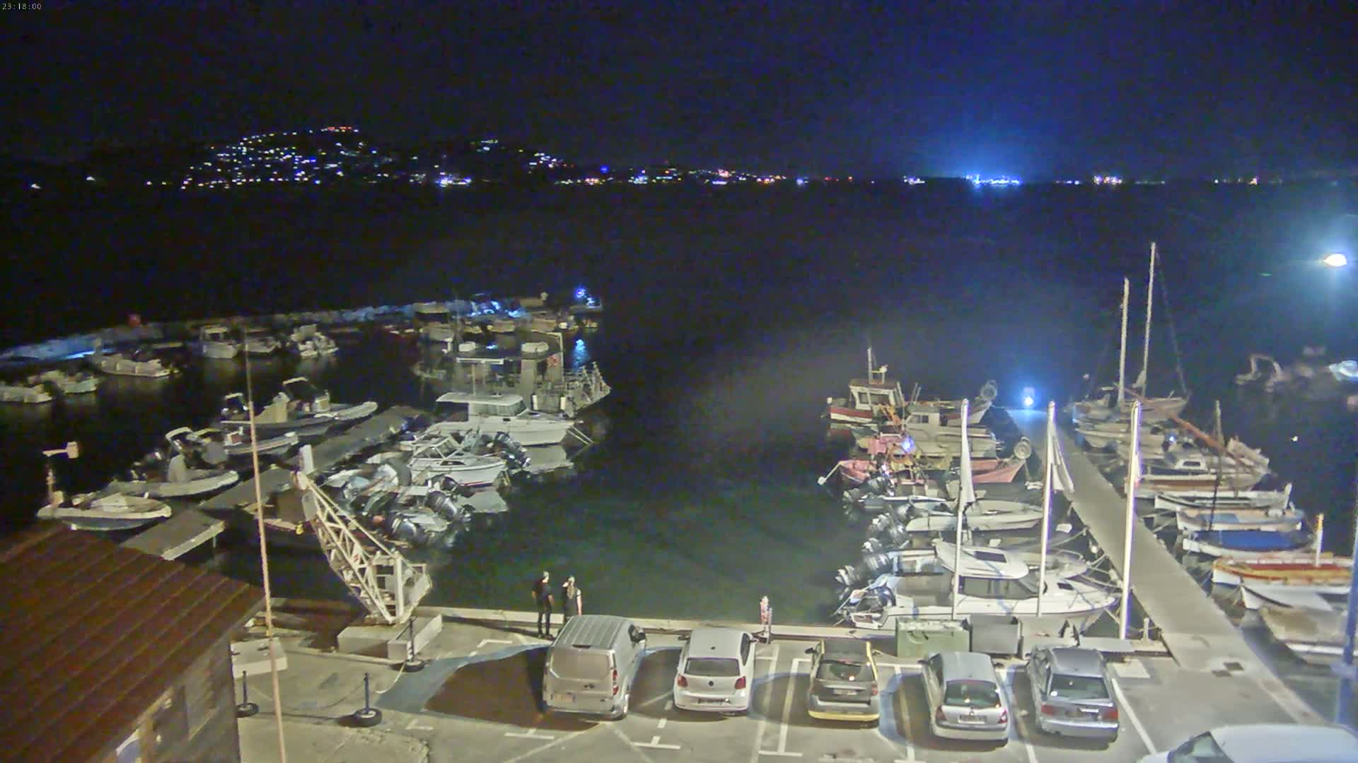 A sunny day at a marina shows numerous boats docked at piers, with additional boats anchored in the calm water beyond.