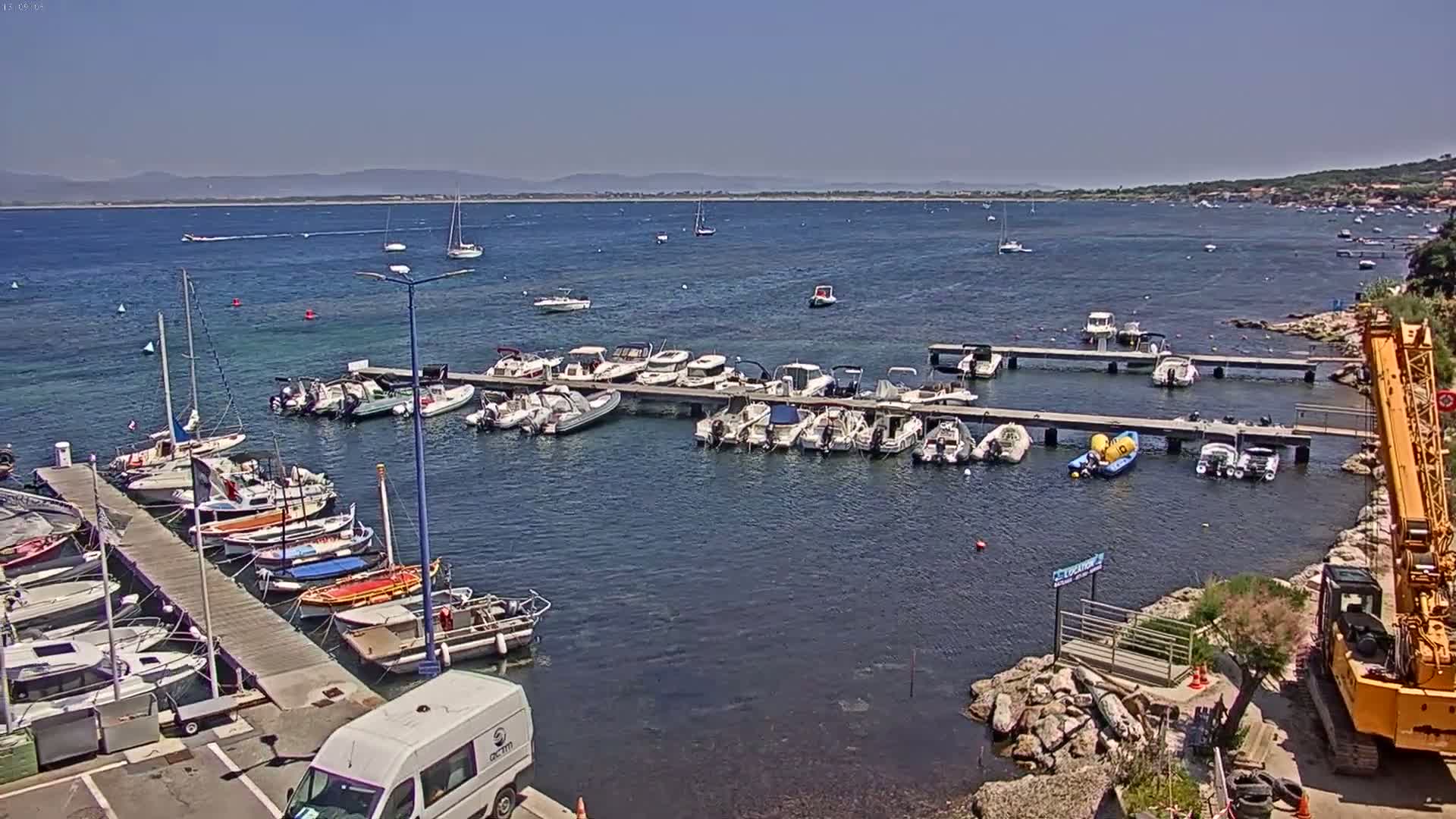 A marina filled with numerous boats of various sizes is nestled in calm, blue water under a clear, sunny sky, with a distant shoreline visible.