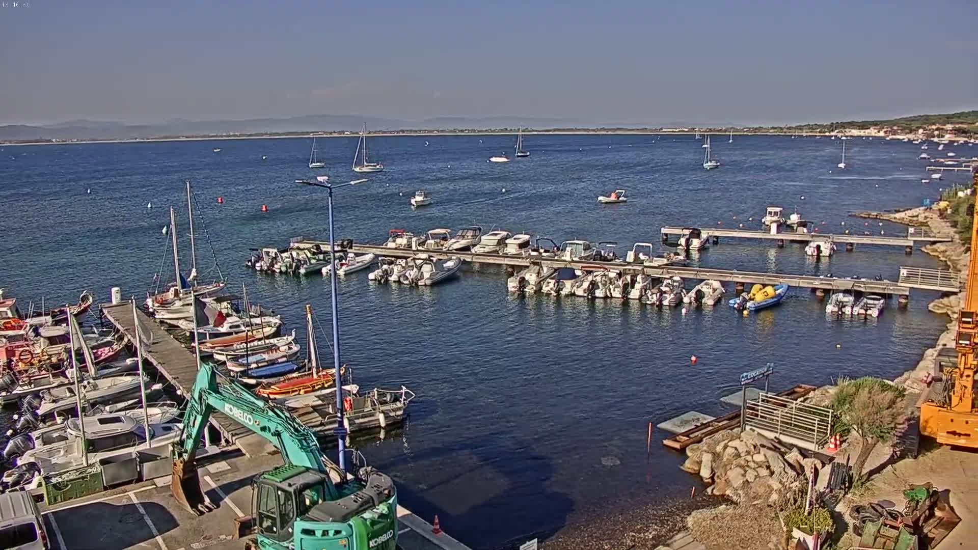A marina filled with numerous boats and yachts on a calm, sunny day, with a clear view of a distant shoreline.