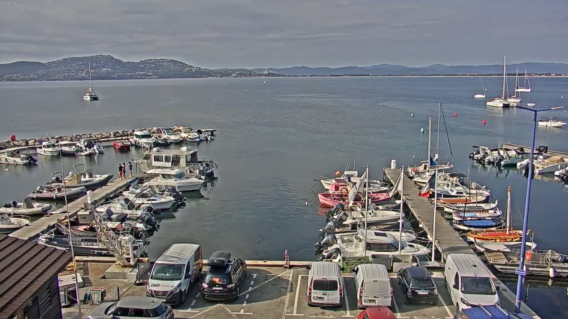 A marina filled with motorboats and sailboats is situated on calm water under a mostly cloudy sky, with distant hills visible in the background.