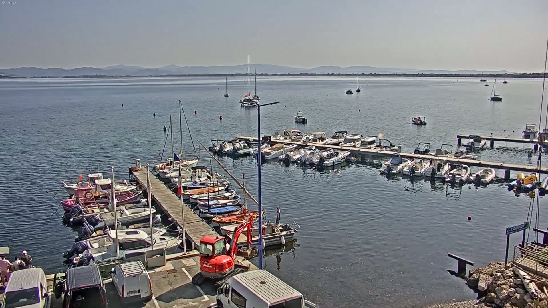 Numerous boats are moored at a marina on a calm, sunny day, with a distant shoreline visible across the water.