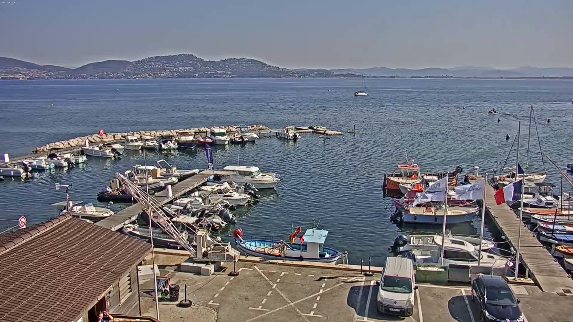 A marina filled with numerous boats of various sizes is situated on calm water under a clear, sunny sky, with distant hills visible on the horizon.