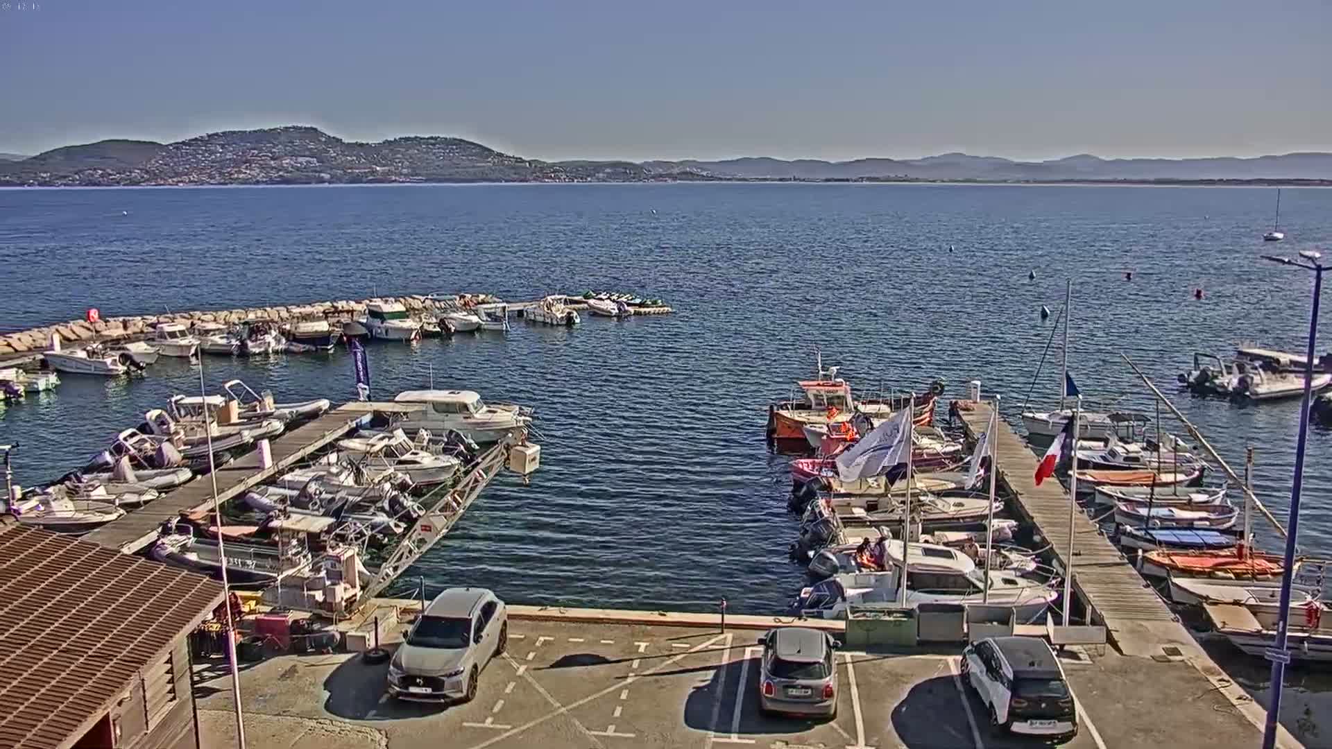 A marina filled with numerous boats of varying sizes is situated near a calm body of water under a clear, sunny sky, with distant hills visible in the background.