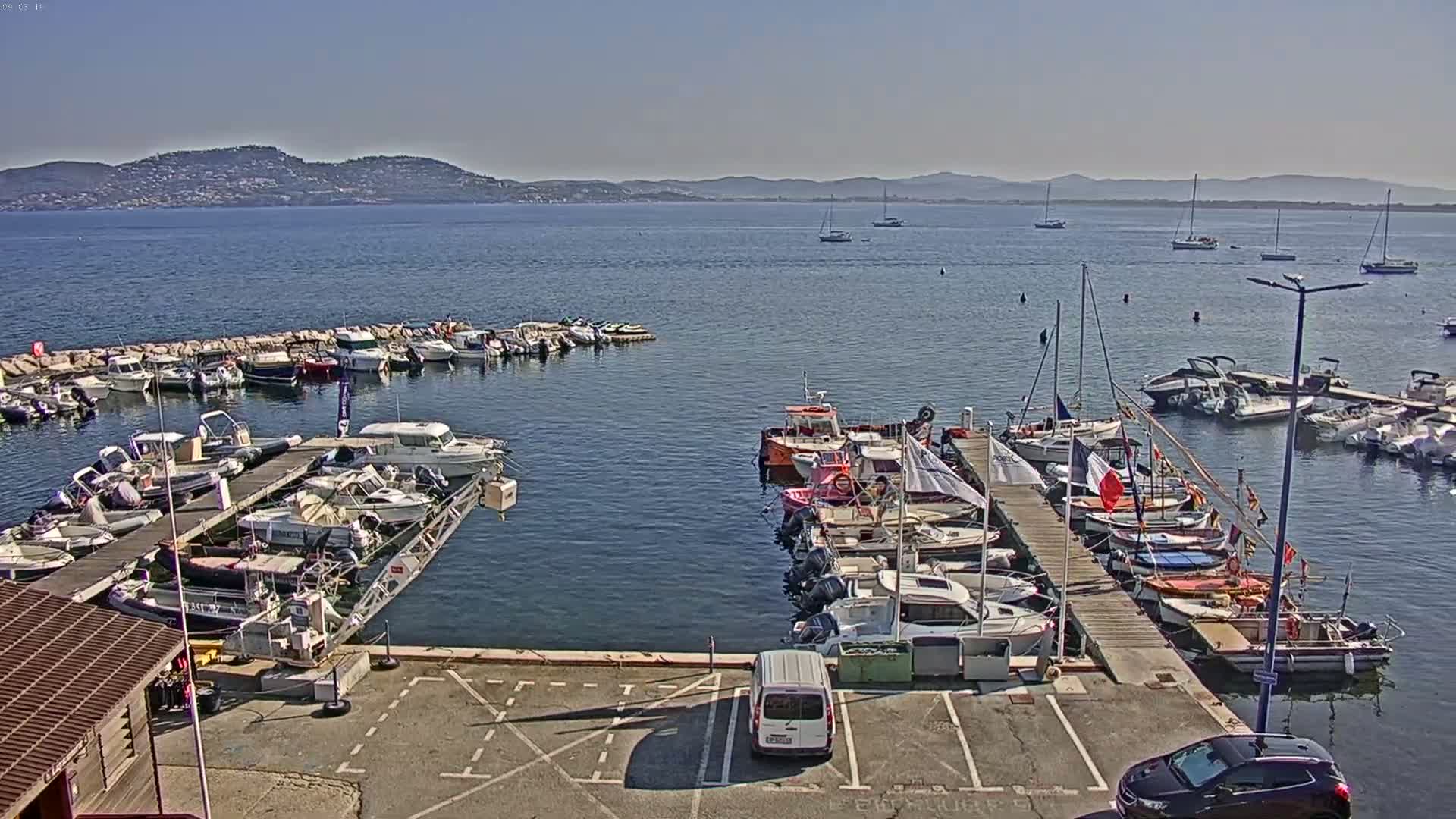 A marina filled with numerous boats and yachts is situated on calm, clear water under a sunny sky, with distant mountains visible on the horizon.