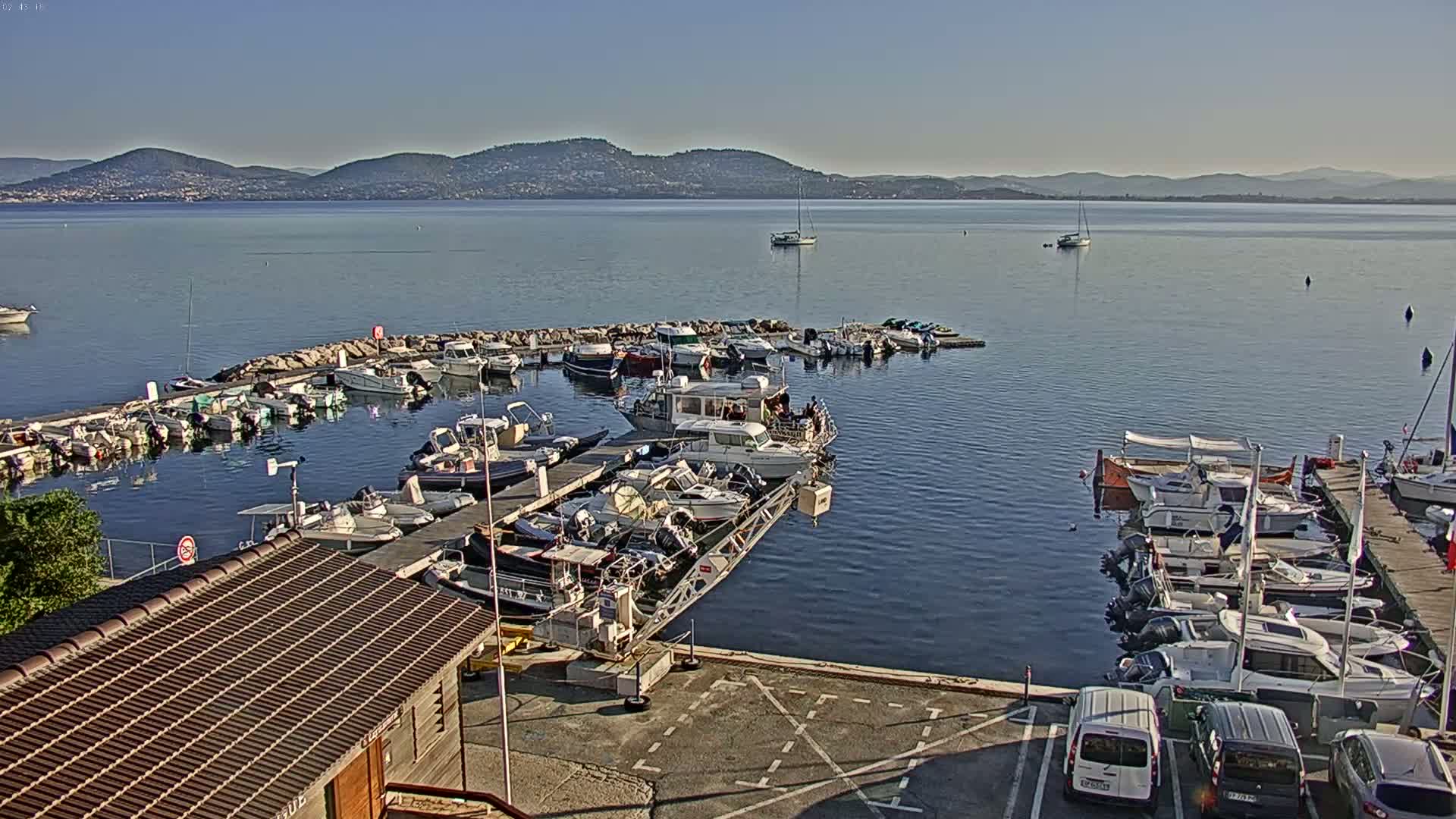 Many boats are docked at a marina on a calm, sunny day with mountains visible in the distance.