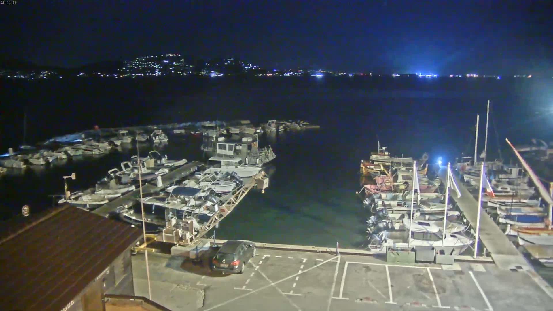 A marina filled with numerous boats at night, under a dark sky, is illuminated by lights from the boats and a distant town across the water.