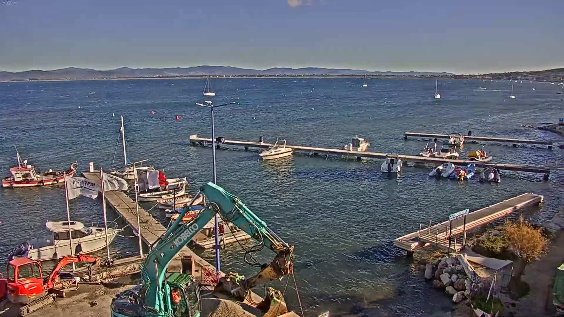 On a clear and sunny day, a bustling bay features numerous boats docked at various wooden piers and anchored in the water, with construction equipment actively working on the gravelly shore against a backdrop of distant mountains.