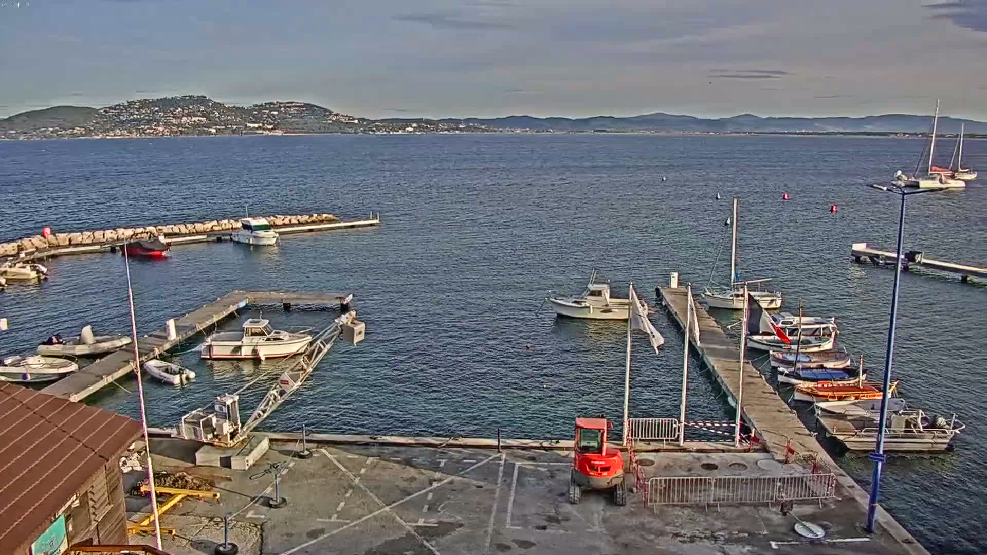 The image shows a tranquil marina filled with various boats docked at wooden piers and a rocky breakwater, with a calm blue sea extending towards distant hills under a partly cloudy sky.