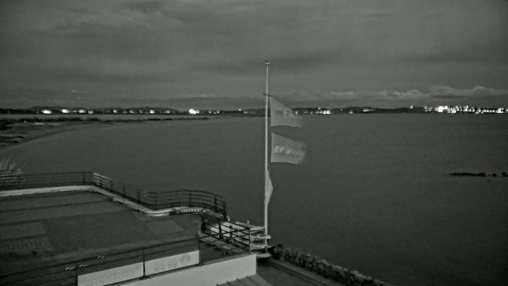 A flagpole with several flags flies at dusk overlooking a calm body of water with a distant shoreline of lights.
