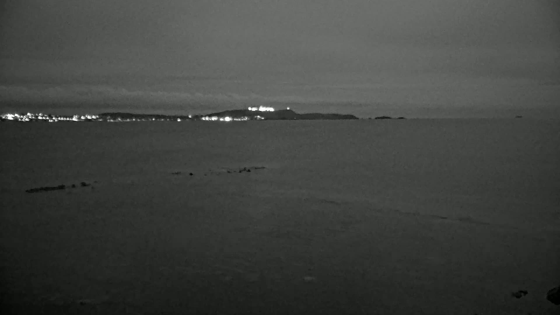 A dark, calm body of water stretches to a distant shoreline illuminated by faint lights under a gray sky.
