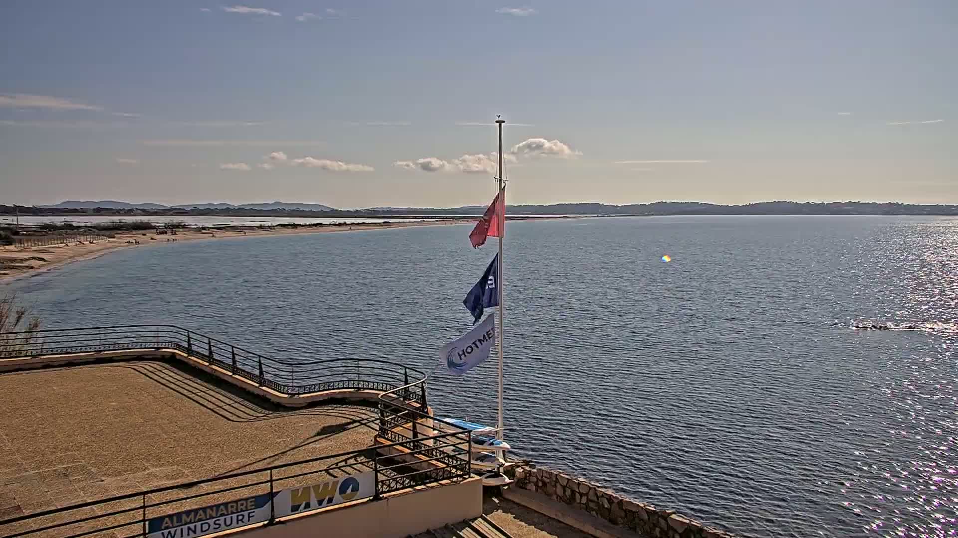 A sandy beach and calm, sunlit body of water are viewed from an elevated position, with a flagpole bearing several flags and a stone structure in the foreground.