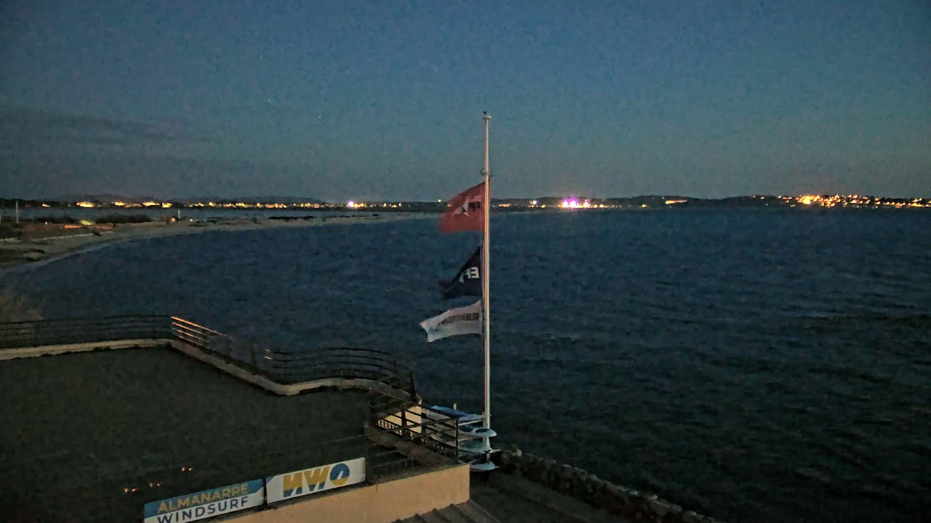A nighttime view of a dark body of water with a distant, illuminated shoreline, a flagpole with three flags, and a flat-roofed building.