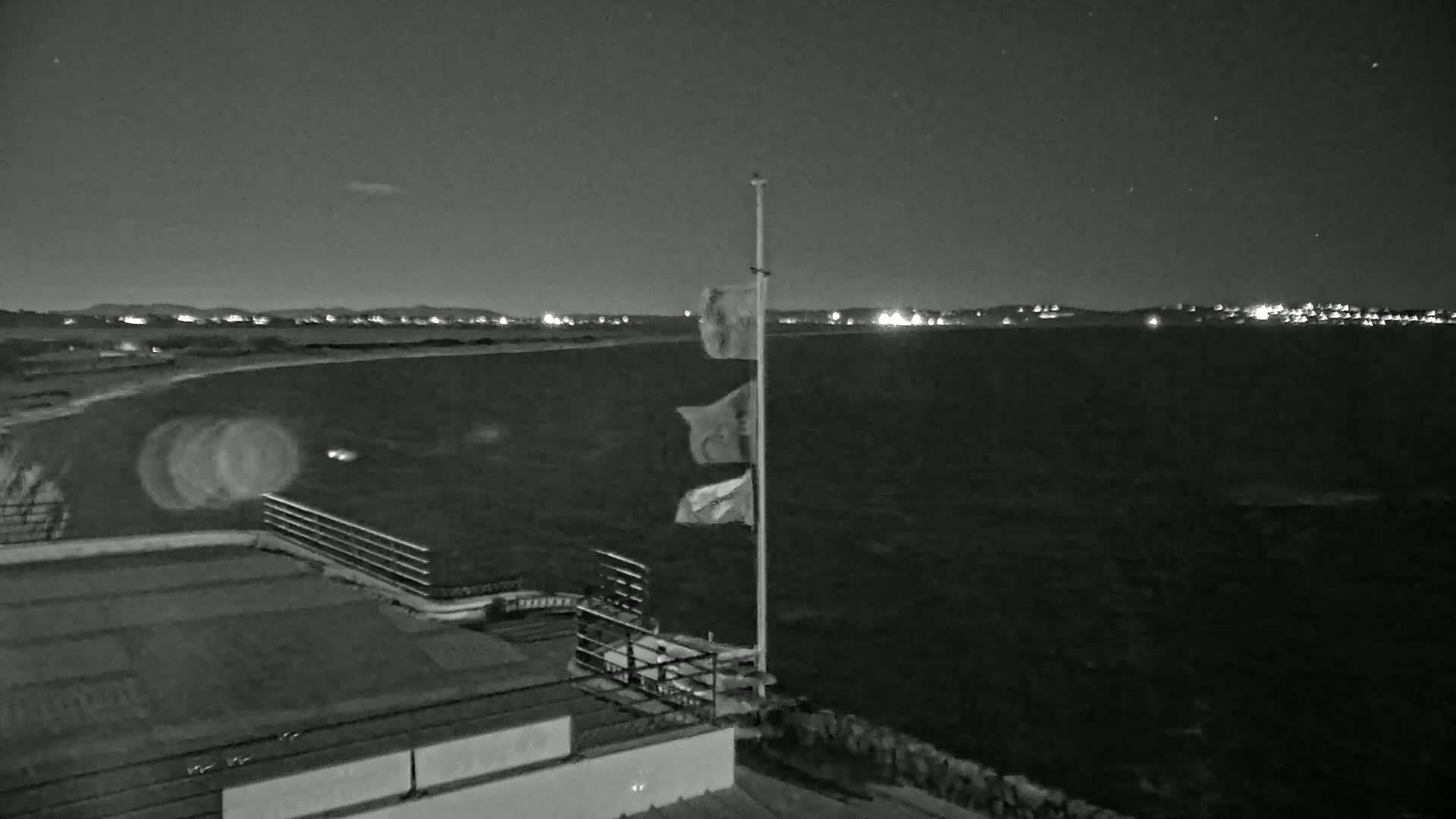 A nighttime view of a dark body of water under a clear, starry sky, with a flagpole bearing several flags near a building's rooftop and distant lights illuminating a shoreline.