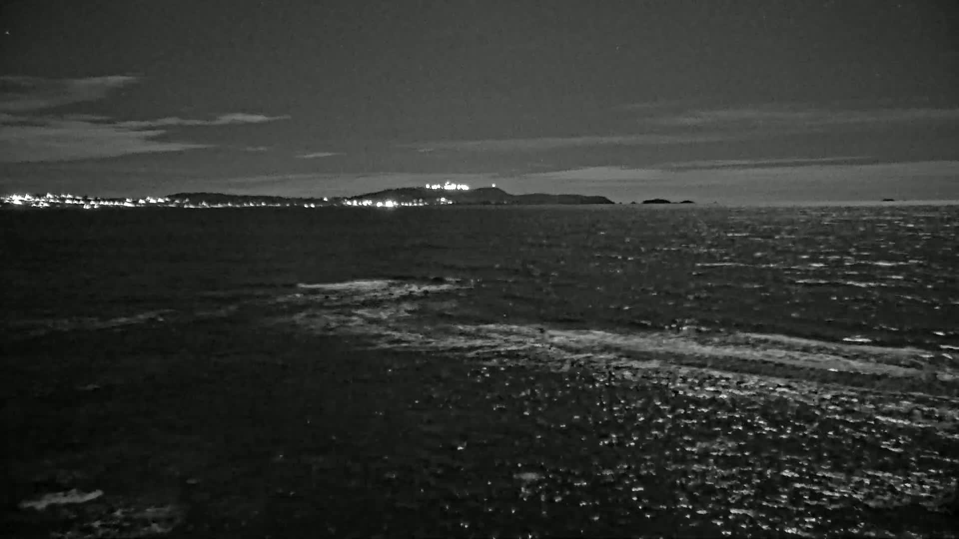 A dark, nighttime seascape shows a distant, faintly lit coastline under a cloudy sky, with a slightly choppy sea in the foreground.