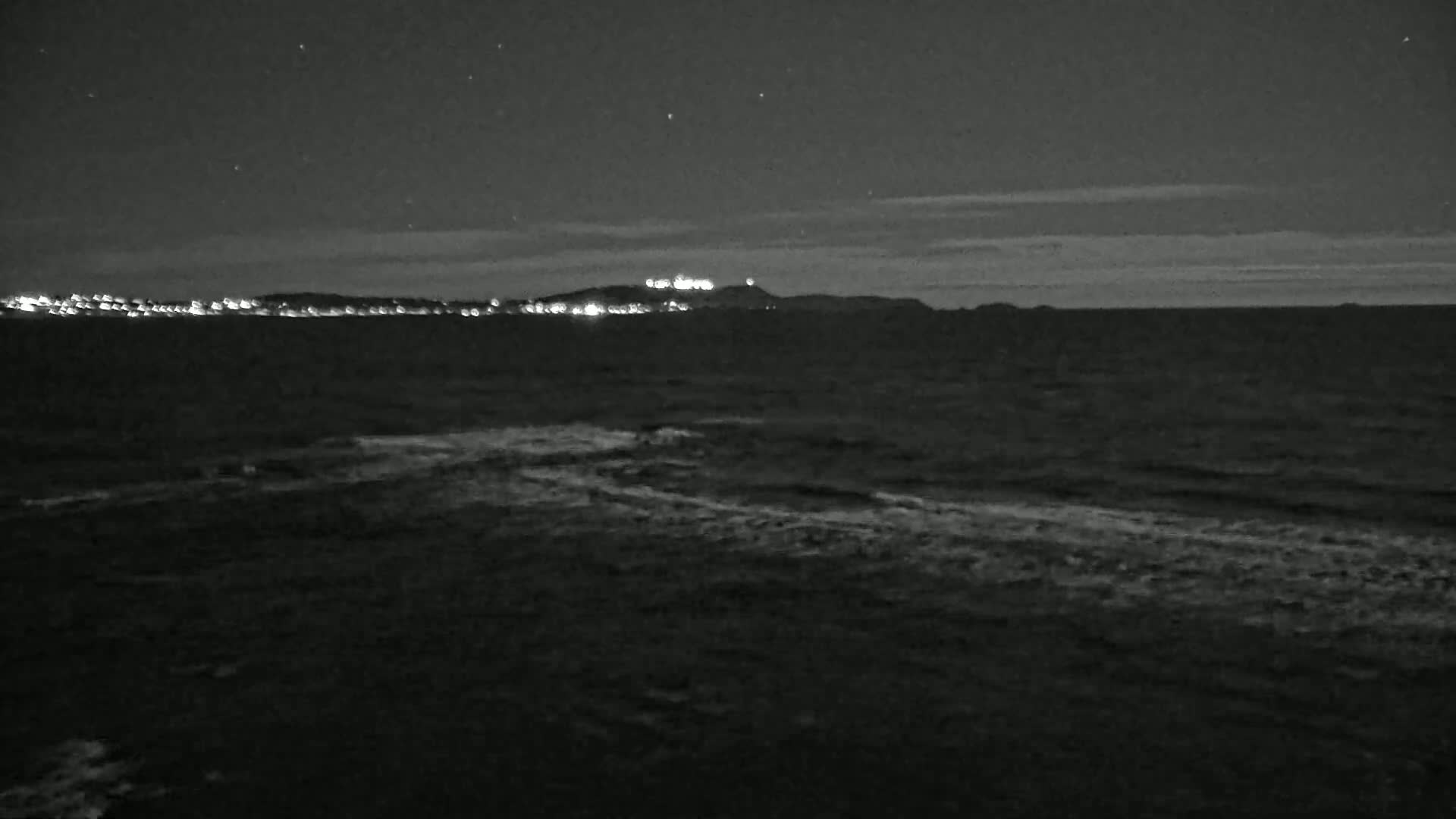 A nighttime view of a dark ocean with a distant shoreline featuring a hilltop with lights under a mostly clear, starry sky.
