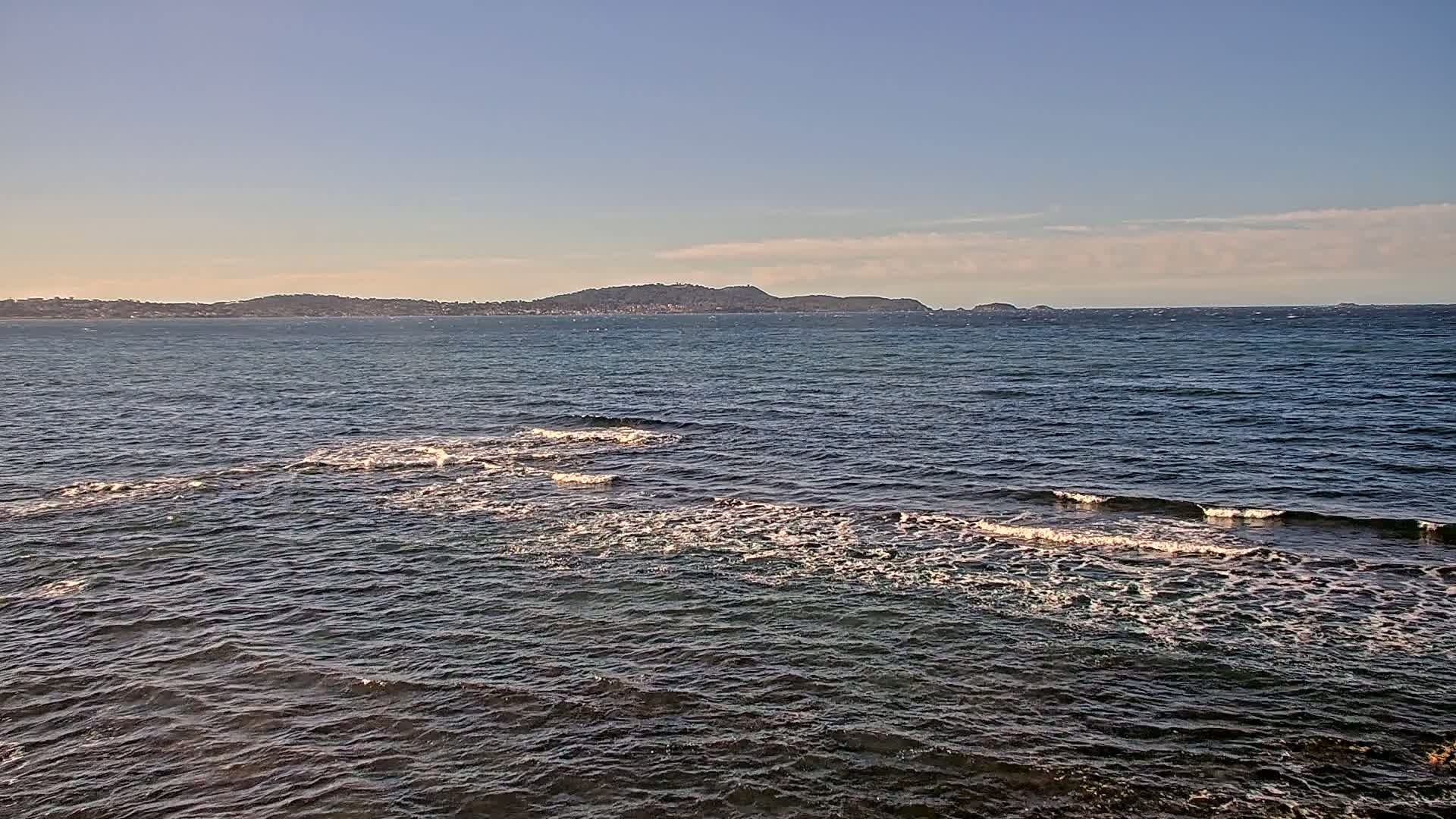A calm sea with small waves laps against a rocky shore, under a clear sky with a distant landmass visible on the horizon.