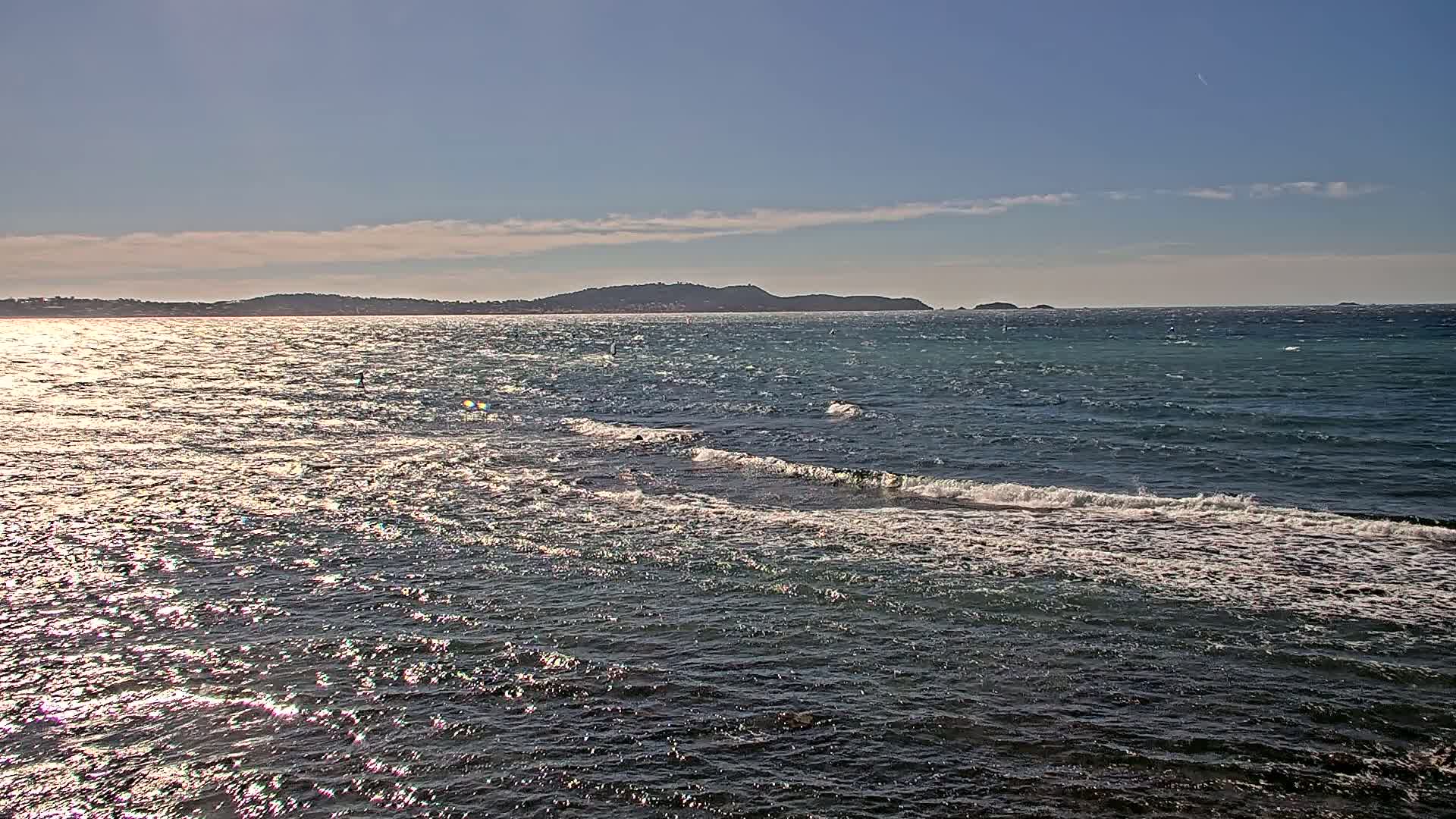 A partly sunny day shows a calm sea with small waves lapping against the shore, a distant landmass visible on the horizon.