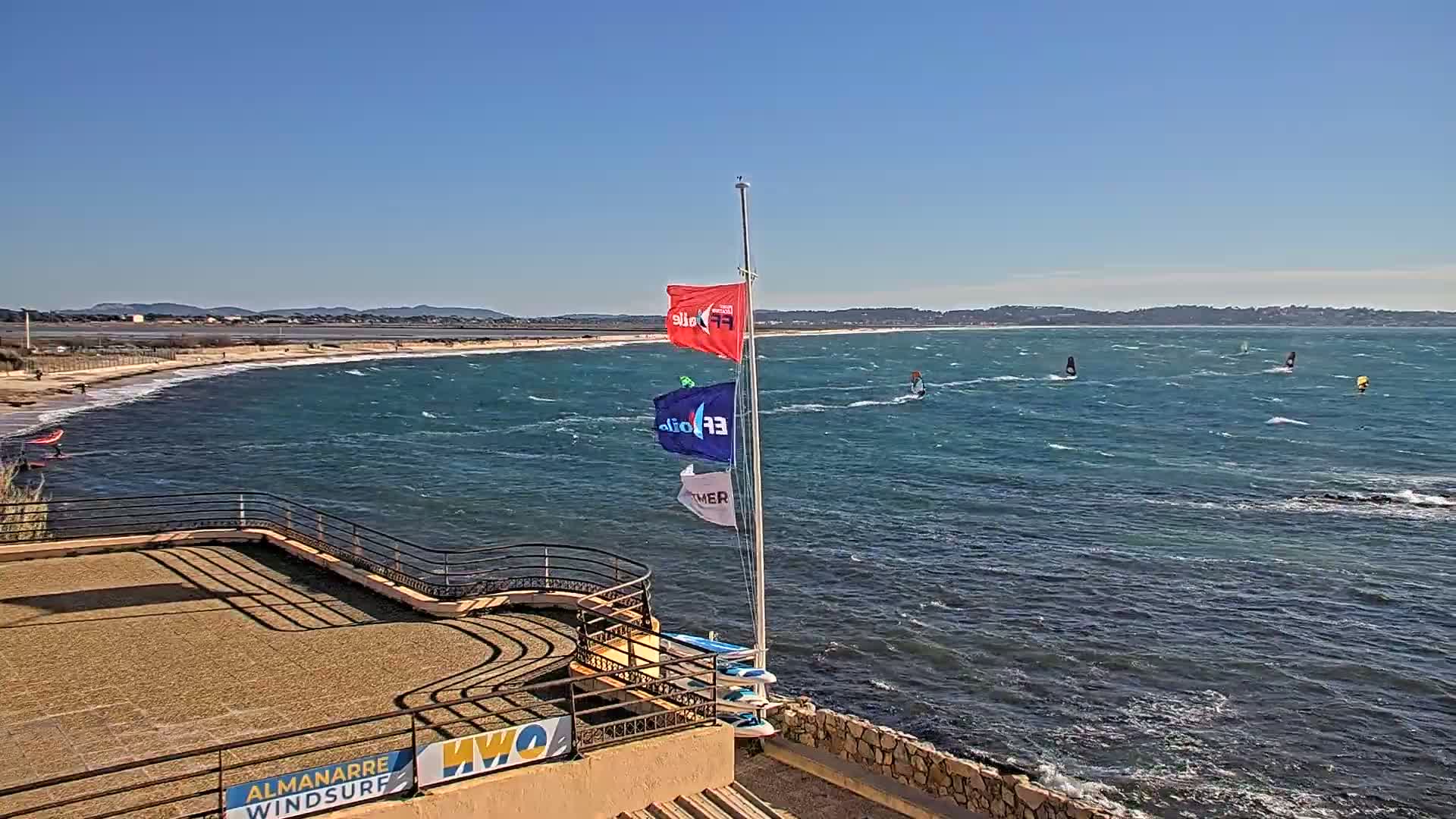 A windy seaside scene shows several windsurfers on a wavy ocean, viewed from an elevated vantage point with a sandy beach in the background.