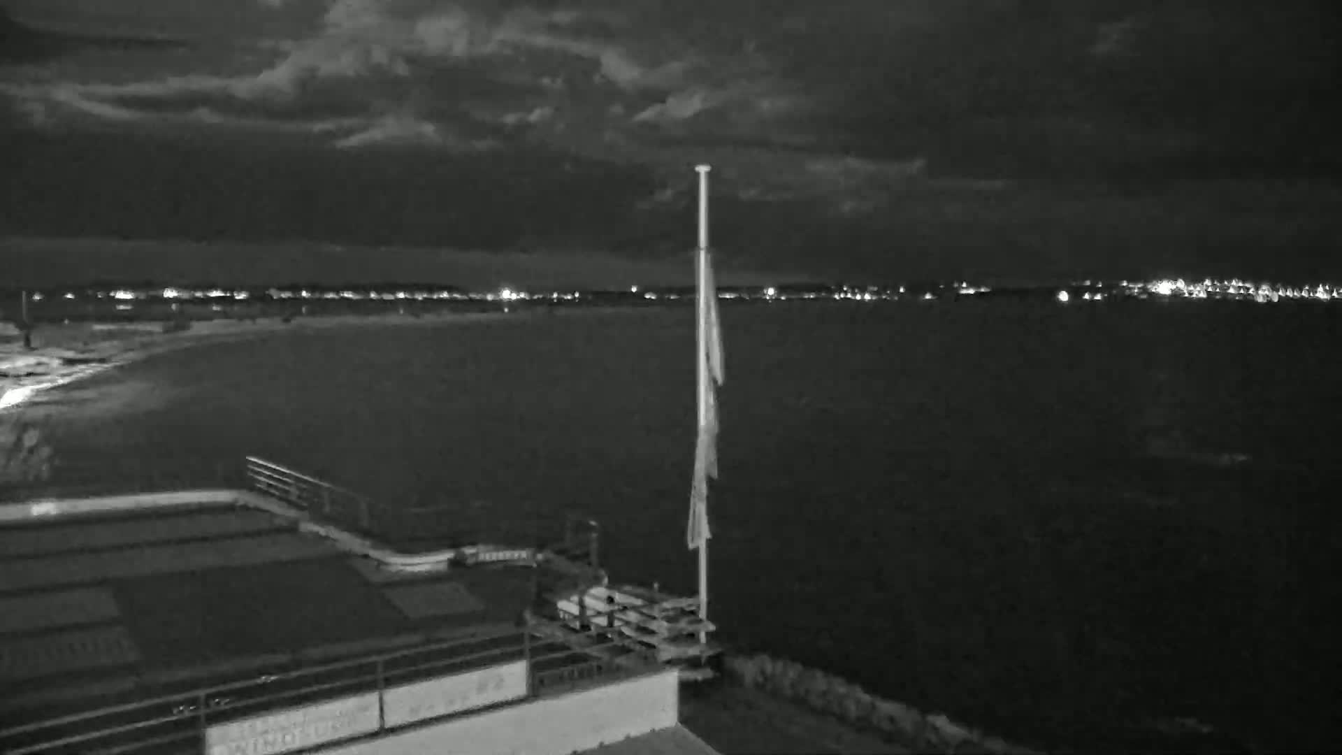 A nighttime view of a body of water with a distant shoreline of lights under a cloudy sky, showing a flagpole and part of a building in the foreground.