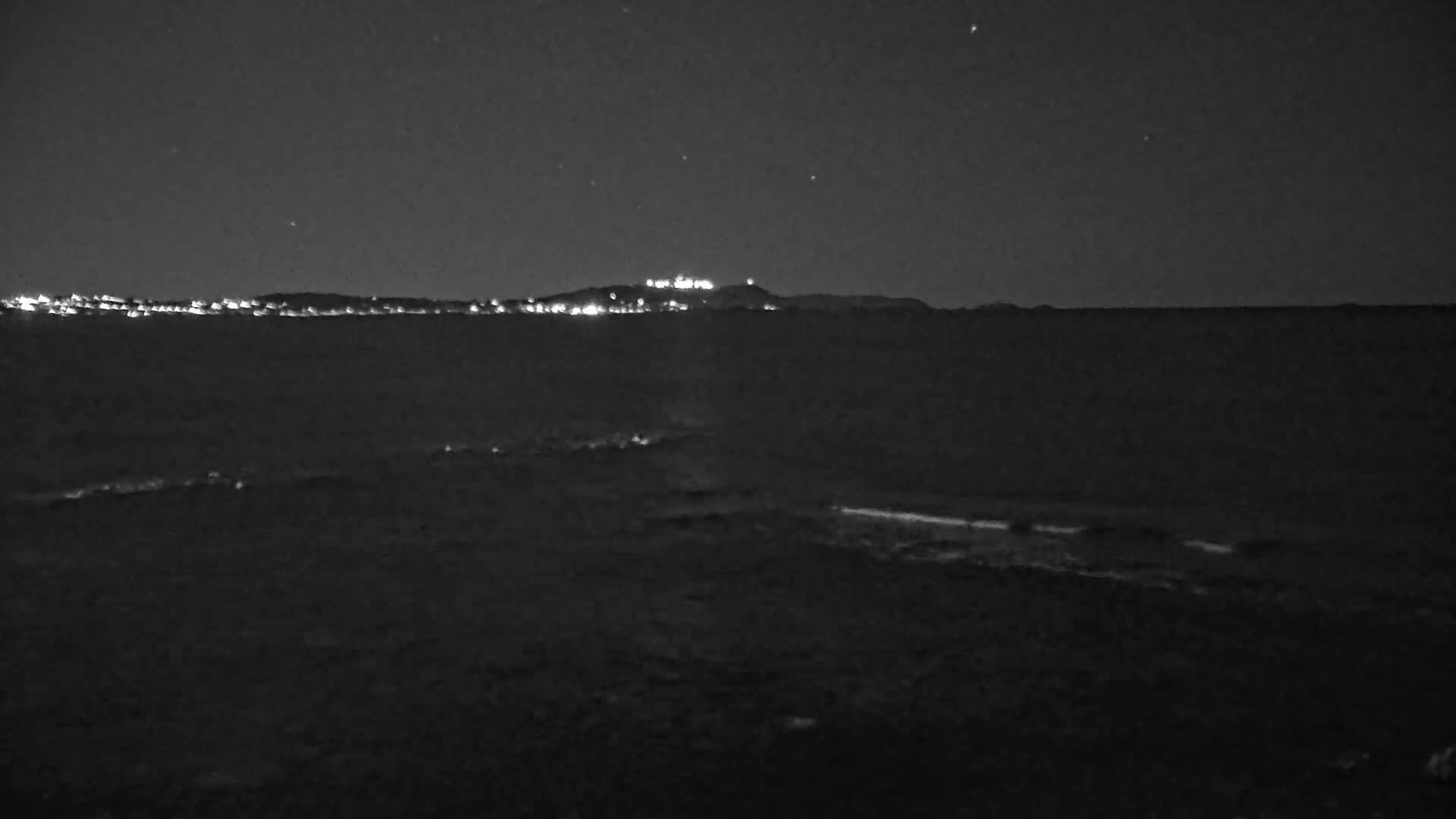 A dark, nighttime coastal scene shows a distant, faintly lit hilltop and shoreline under a clear, starry sky.