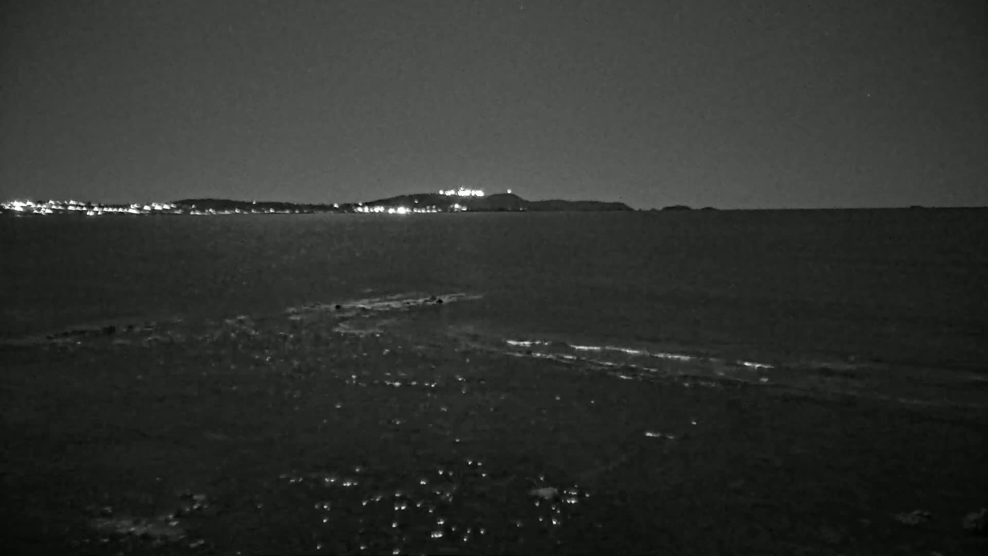 A dark, nighttime scene shows a calm body of water with a distant shoreline featuring illuminated buildings and a hilltop.